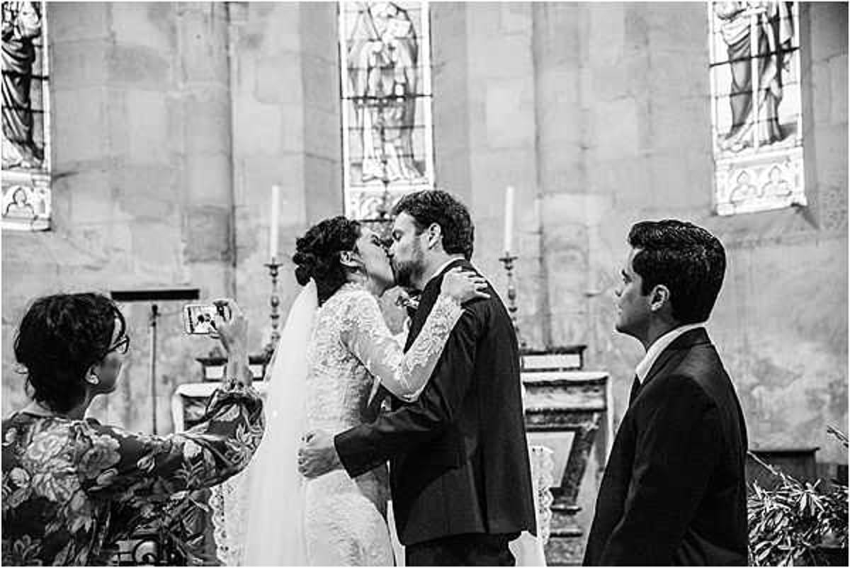 Black-and-white image of the first kiss moment during a church wedding ceremony. The bride, wearing a long-sleeved lace gown with her dark hair pinned up, and the groom, in a dark suit, share a kiss at the altar, with stained glass windows and altar candles visible in the background. To the left, a female guest photographs the moment on a smartphone, while a male guest in a dark suit stands to the right observing. The shot is a medium wide frame capturing the couple and two surrounding guests, with high contrast tones emphasizing the stone church interior.