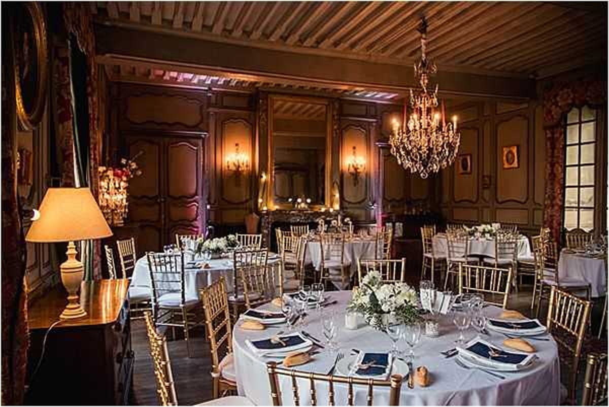 A wide interior shot of a chateau ballroom set up for a wedding reception dinner. The room features wood-paneled walls with warm amber uplighting, a crystal chandelier hanging from a painted coffered ceiling, and wall sconces providing soft ambient light. Multiple round tables are dressed in white floor-length linens and set with navy blue folded napkins, gold charger plates, and glassware, surrounded by gold chiavari chairs. Centerpieces consist of low arrangements of white flowers, likely white roses or hydrangeas, with greenery. A table lamp with a cream shade is visible on a sideboard to the left, and red floral arrangements add accent color near the entrance. The overall decor palette combines navy, gold, and white against the warm-toned paneled interior, reflecting a classic French chateau style. Potential venue feature image.