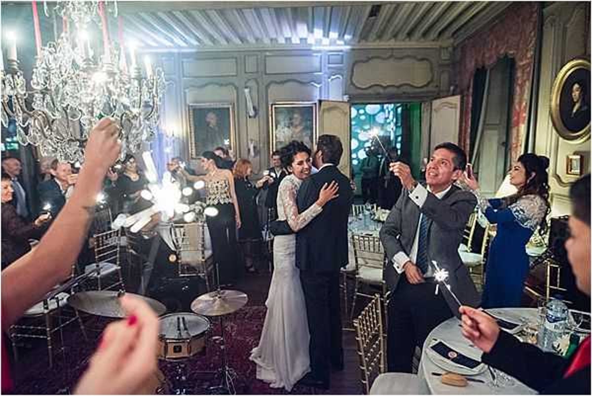 The bride and groom share their first dance in an ornate chateau ballroom while surrounding guests hold lit sparklers. The bride wears a form-fitting white gown with lace long sleeves, and the groom is in a black suit. Approximately 20–30 guests line the dance floor, with gold chiavari chairs and formally set dining tables visible in the background. The room features wood-paneled walls, crystal chandeliers, oil portrait paintings, and a live drum kit is visible to the left, indicating a live band performance. The warm sparkler light contrasts with the ambient room lighting in this wide-angle reception shot.