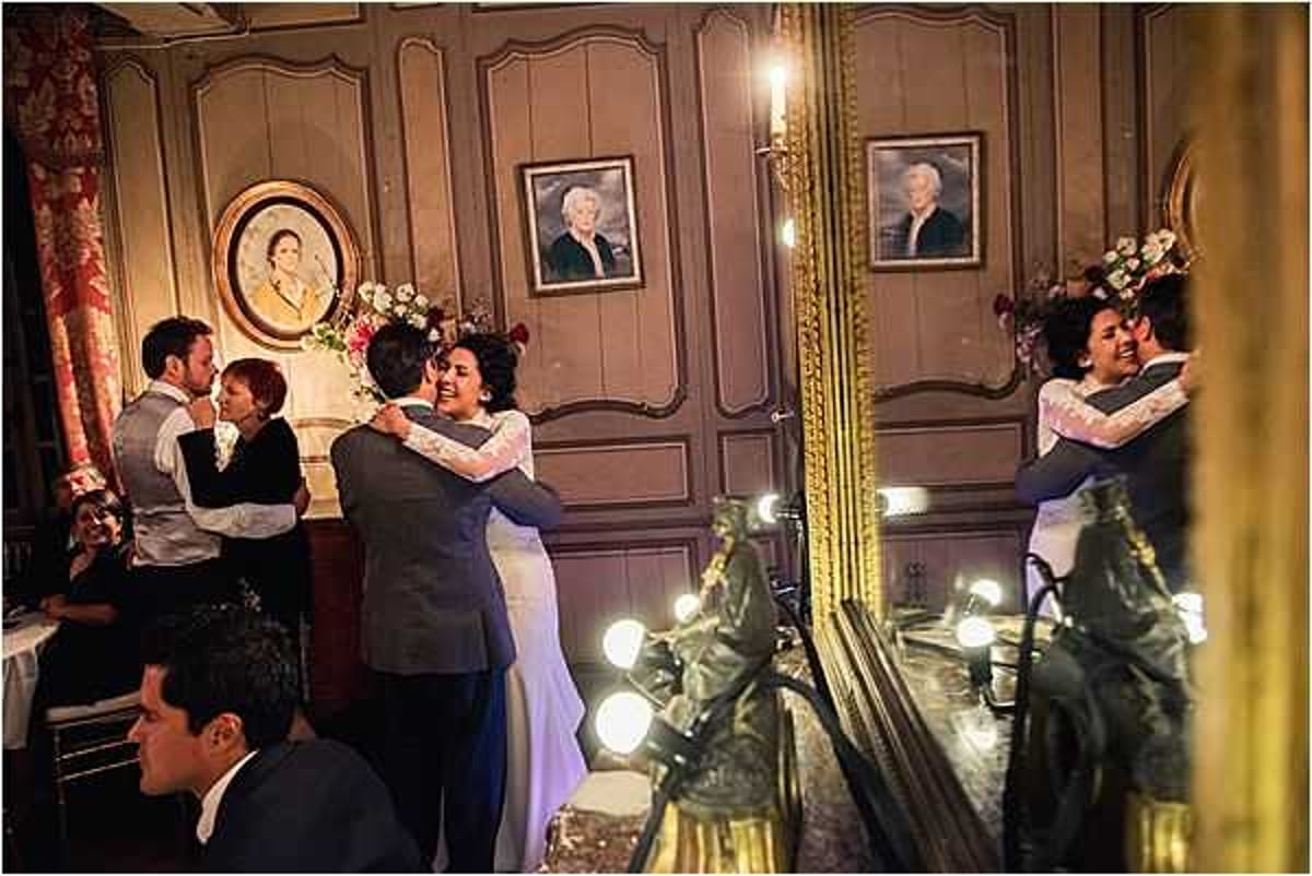 A first dance scene captured indoors in what appears to be a classic French chateau or manor room, with wood-paneled walls painted in a warm taupe and framed portrait paintings hanging on the wall. The bride wears a white gown with lace long sleeves and the groom is dressed in a dark navy suit; they are embracing and smiling during their first dance. A large ornate gold-framed mirror to the right reflects the couple, creating a dual view of the moment, while warm Edison-style bulb lighting along the mirror frame adds a soft glow. In the background, two other guests — one in a light grey suit — are also embracing or dancing, and a seated male guest in a dark suit is visible in the foreground.