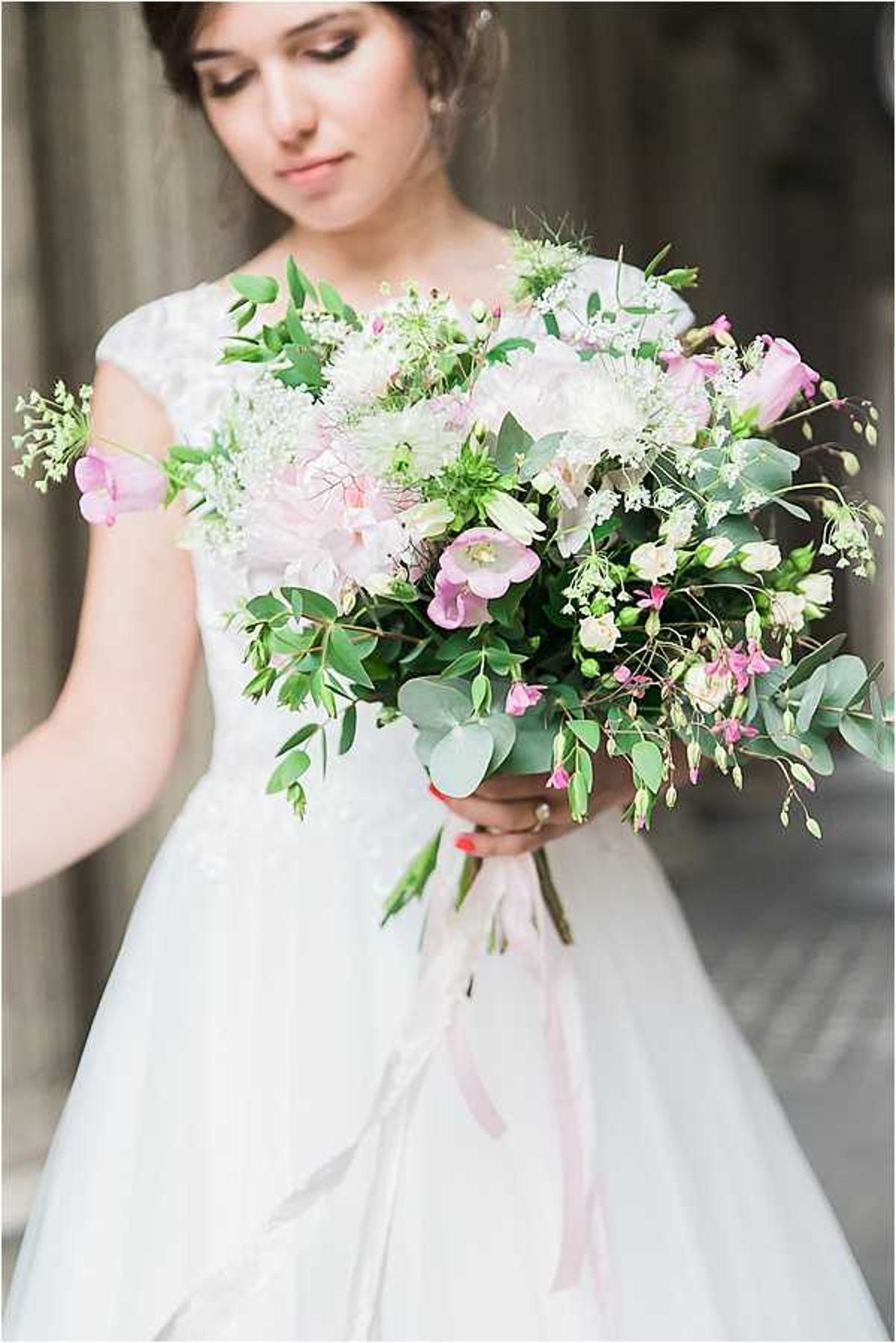 A close-up portrait of a bride looking down at her bouquet, set against a softly blurred architectural background. She wears a white cap-sleeve lace bodice dress with a tulle skirt, with her dark hair pulled up. The bouquet is a loose, garden-style arrangement featuring blush peonies, pink lisianthus, white Queen Anne's lace, cream freesia buds, eucalyptus, and trailing greenery, tied with a long blush pink ribbon. The composition is a medium portrait shot with the bouquet in sharp focus in the foreground and the bride's face softly focused in the background.