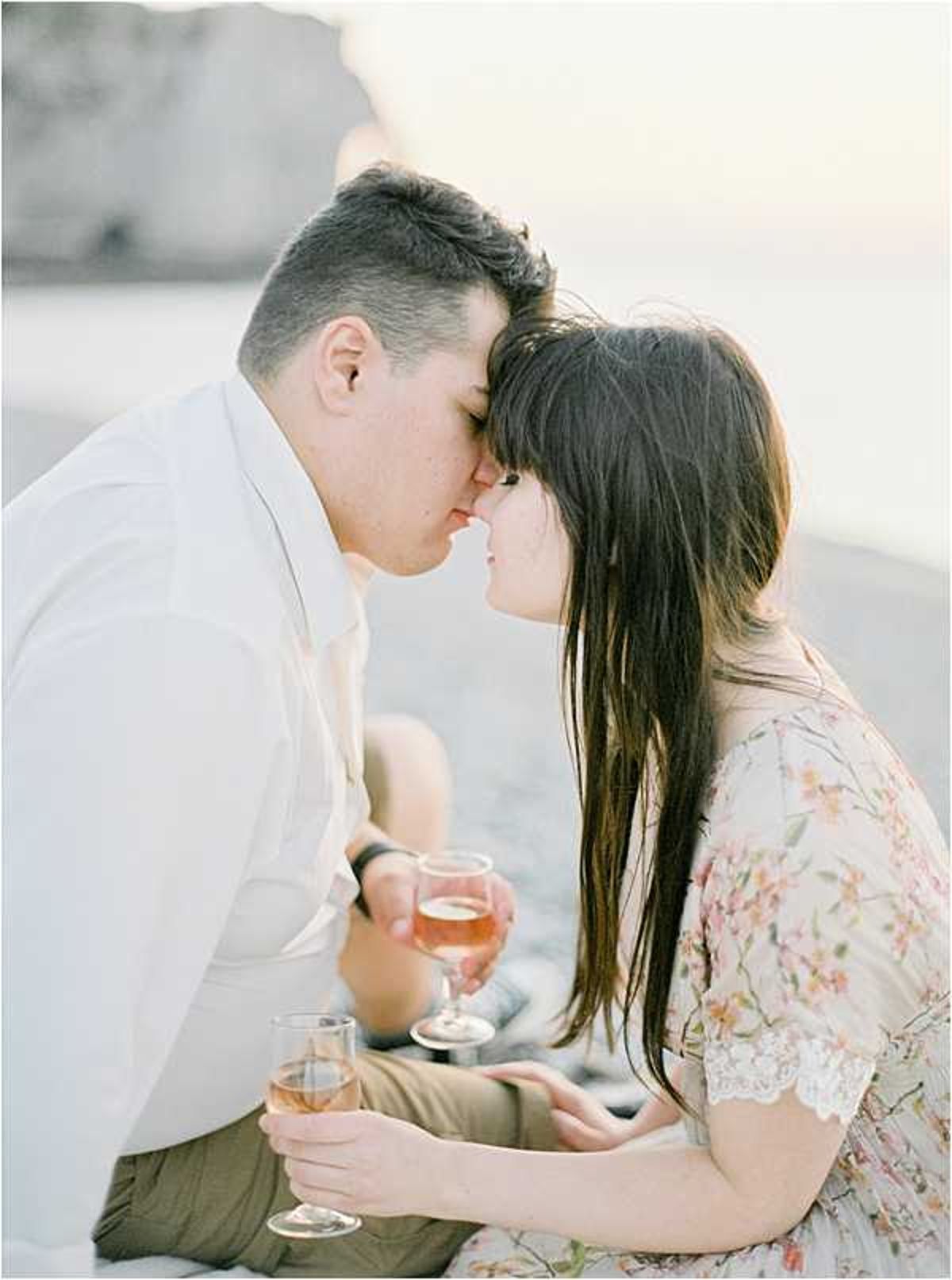 A couple shares a close, near-kiss moment during what appears to be an engagement or pre-wedding portrait session outdoors. They are seated together, each holding a glass of rosé wine, with a coastal cliff visible softly out of focus in the background. The man wears a white button-down shirt with khaki shorts, while the woman wears a light floral dress with a lace trim detail featuring small pink and green blossoms. The image is shot in a soft, airy film style with warm, muted tones and shallow depth of field, captured as a close-up portrait from the side.