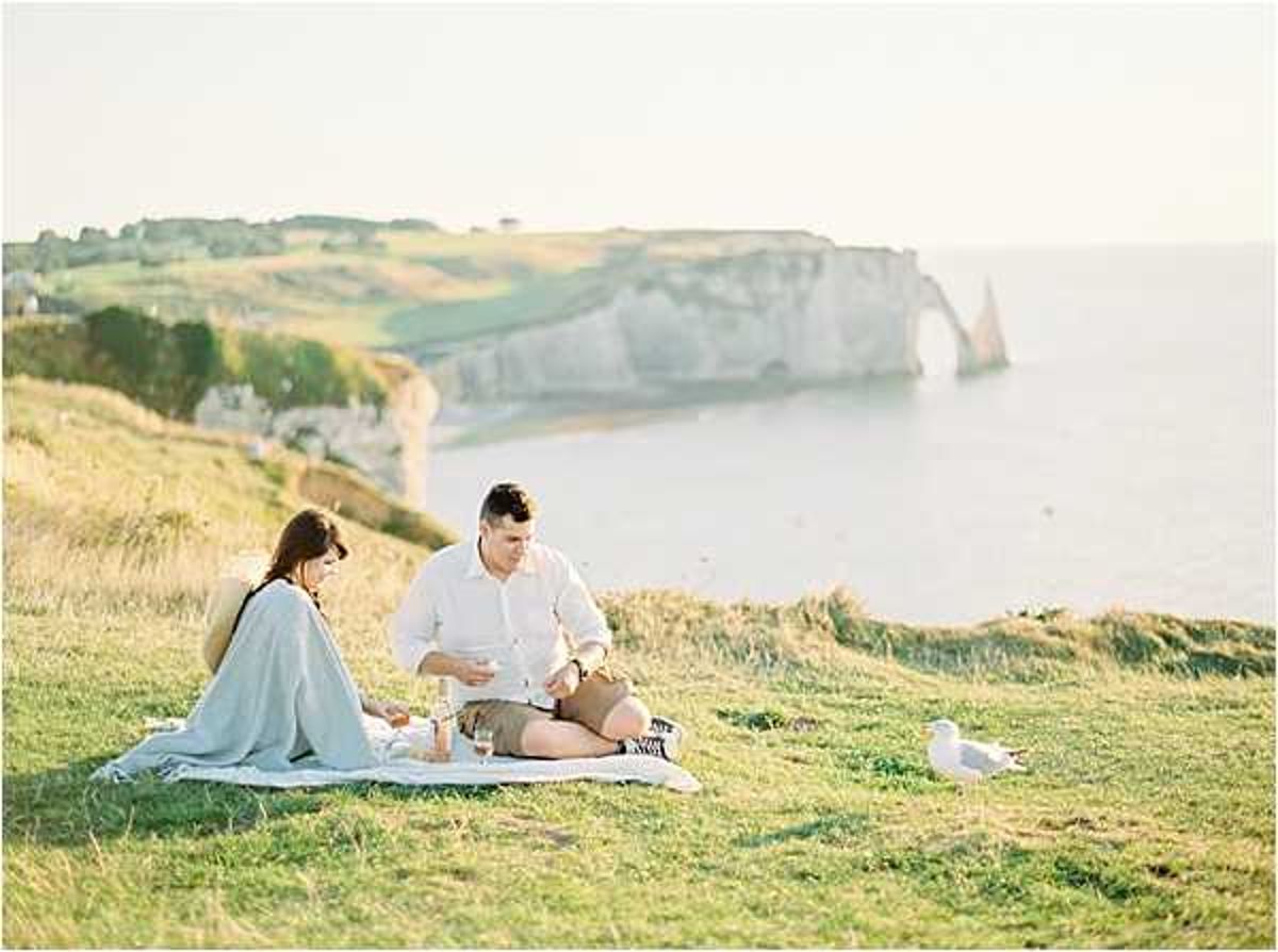An engagement or couples portrait session featuring two people seated on a white blanket set up as a picnic on a clifftop overlooking the iconic chalk cliffs and natural arch of Étretat in Normandy, France. The woman wears a light blue wrap or shawl and the man wears a white linen shirt and shorts, and they are looking at something together, with wine glasses visible on the blanket. A seagull stands nearby on the grass, and the scene is lit by warm golden hour light. The composition is a wide environmental shot that places the couple small within the dramatic coastal landscape.