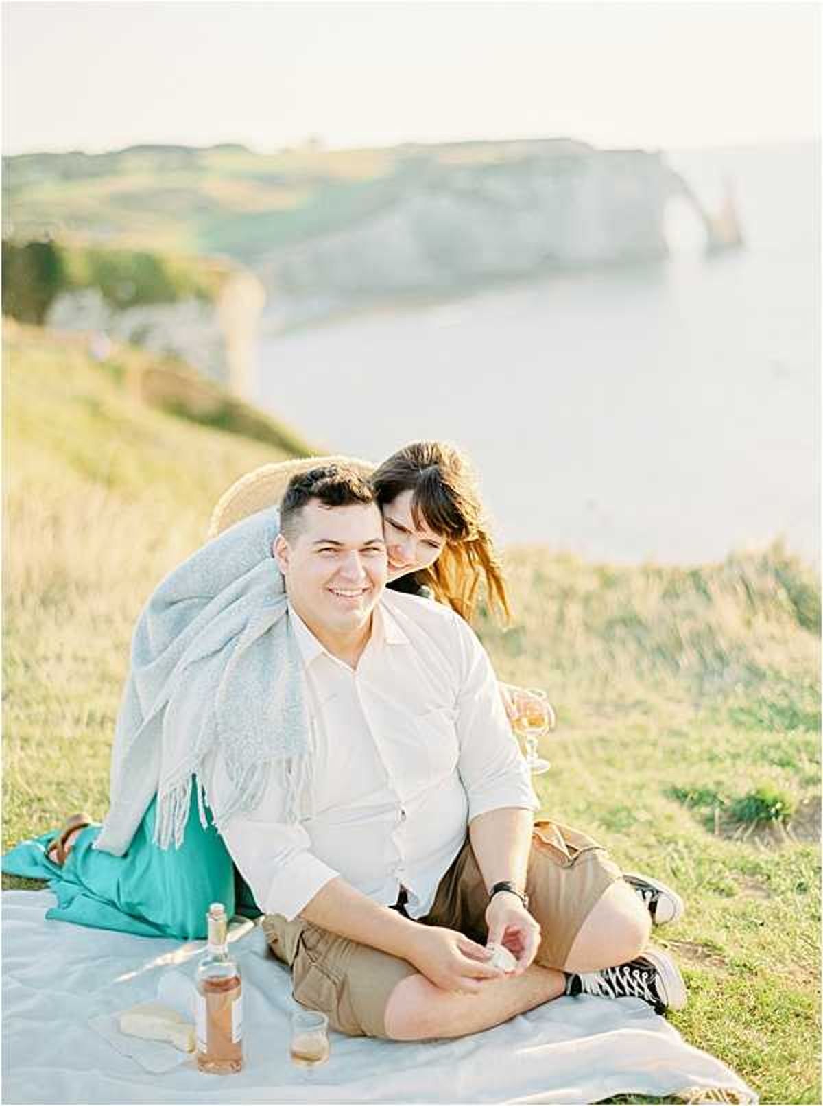 A couple sits together on a white blanket on a clifftop overlooking the sea, in what appears to be an engagement or pre-wedding portrait session at the cliffs of Étretat in Normandy, France. The man wears a white button-down shirt and khaki shorts, while the woman wears a teal skirt and has a light grey fringed blanket draped over her shoulders; she leans affectionately against him from behind. A bottle of rosé wine, a glass, and what appears to be a small cheese or snack are arranged on the blanket, styling the scene as a casual clifftop picnic. The shot is a medium portrait taken at golden hour with warm, soft backlighting, and the iconic chalk arch rock formation is visible in the soft-focus background.