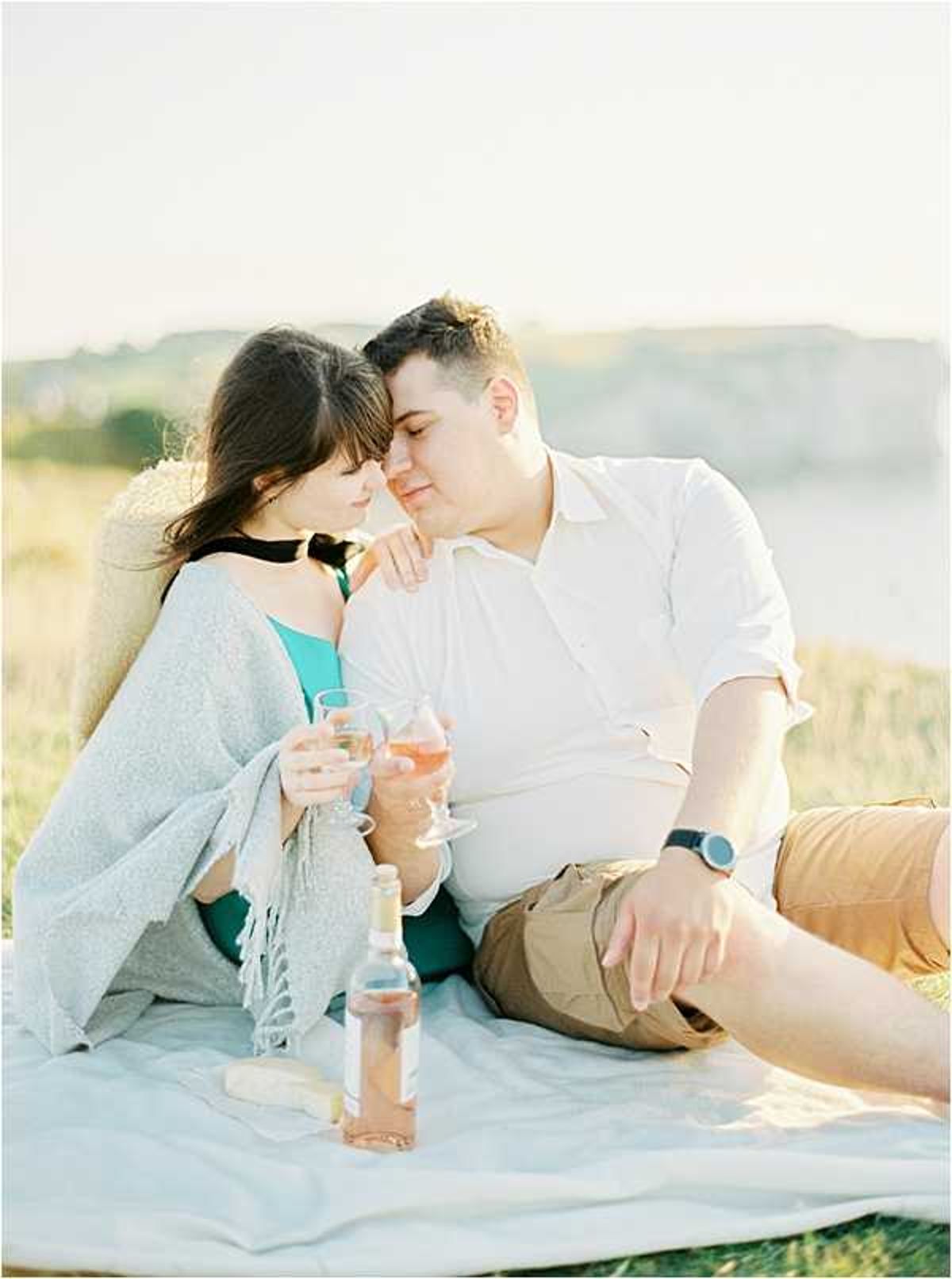 An engagement or couples portrait session shot outdoors, with a man and woman seated closely together on a white blanket in an open landscape setting with a coastal or hillside backdrop. The woman wears a teal top with a light grey fringed wrap and a black choker necklace, while the man wears a white short-sleeve button-down shirt and khaki shorts with a dark watch. Both hold glasses of rosé wine, and a bottle of rosé is placed on the blanket in front of them. The couple is leaning into each other with foreheads nearly touching in an intimate moment. The image is shot in warm, soft golden-hour light with a slightly overexposed, film-style aesthetic. Medium portrait shot.