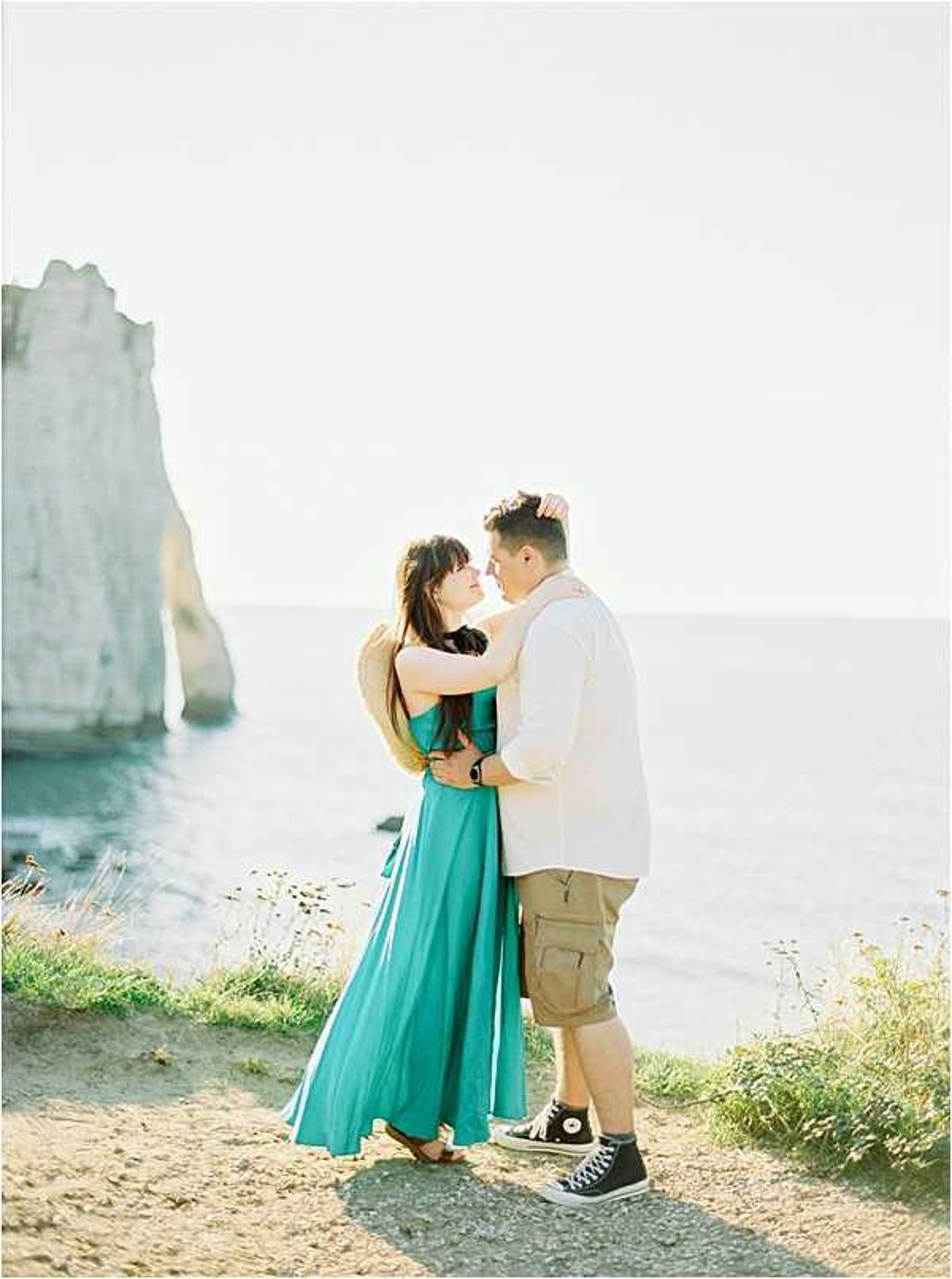 A couple portrait shot outdoors on a clifftop overlooking the sea, with the iconic white chalk arch cliffs of Étretat visible in the background. The woman wears a teal/turquoise sleeveless maxi dress and holds a straw hat, while the man wears a white linen shirt, khaki cargo shorts, and black Converse high-top sneakers. The two are facing each other in a close embrace, foreheads nearly touching, in a relaxed and casual style. The image is a medium full-body shot with soft, bright natural light giving a slightly overexposed, airy film-like quality.