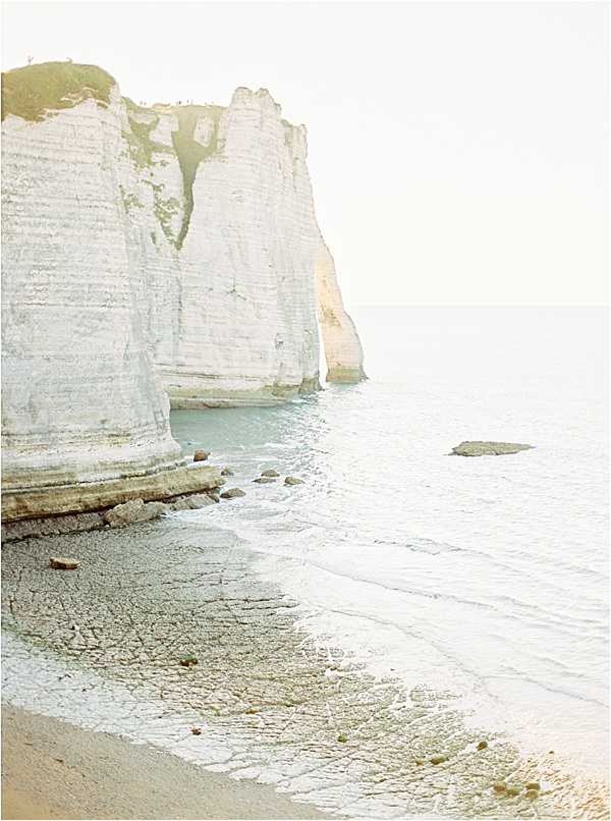 This image contains no people, wedding party, or wedding-related activity. It is a wide landscape shot of white chalk cliffs meeting a calm shoreline, likely the cliffs of Étretat in Normandy, France. The image has a soft, washed-out film quality with pale blue-green water and a sandy pebble beach in the foreground. This appears to be a location or venue context shot rather than a wedding photograph.