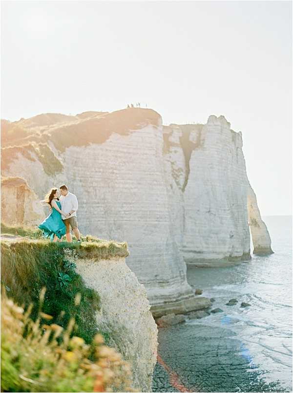 A couple poses together on the edge of white chalk cliffs overlooking the sea, in what appears to be an engagement or pre-wedding portrait session at Étretat in Normandy, France. The woman wears a flowing teal/turquoise dress that billows in the wind, while the man is dressed in a casual white shirt and khaki trousers. They stand close together facing each other against a backdrop of dramatic chalk cliff formations and ocean water below. The wide-angle shot captures the full scale of the coastal landscape, with warm golden backlight creating a sun-flare effect across the scene.