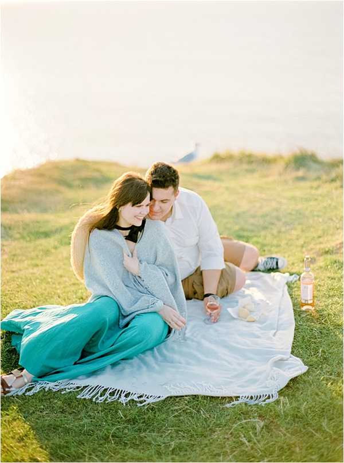 A couple sits together on a white fringed blanket during an outdoor engagement or couples portrait session on a coastal grassy clifftop at golden hour. The woman wears teal wide-leg trousers and a grey knit wrap, while the man is dressed in a white shirt and khaki shorts; they are seated close together, smiling and holding wine glasses, with a bottle of rosé and what appears to be bread or cheese also on the blanket. The composition is a medium shot with warm, soft golden-hour light bathing the scene. The styling is casual and relaxed with a picnic setup.