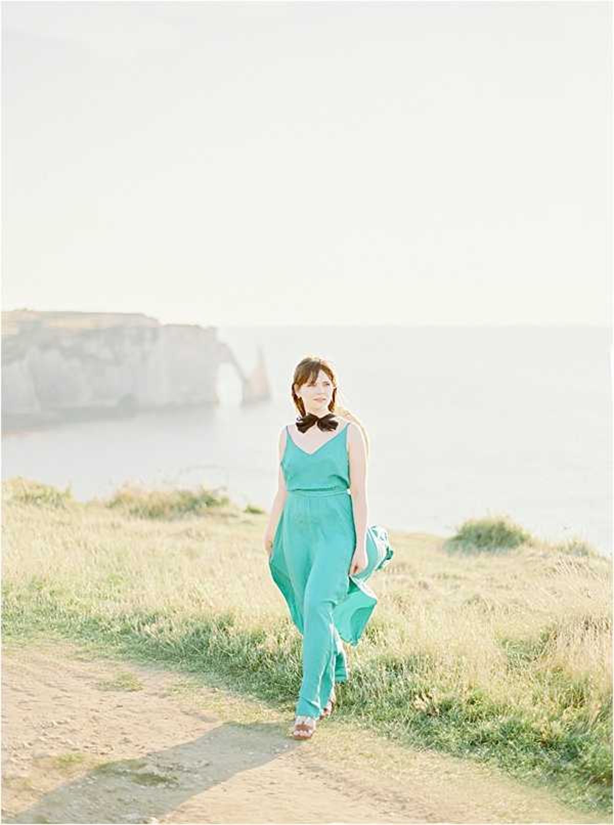 A solo portrait of a woman walking along a clifftop path with the iconic chalk cliffs and natural arch of Étretat visible in the background over the sea. She is wearing a teal/turquoise sleeveless jumpsuit with a black bow tied at the neckline, and flat sandals, with fabric catching slightly in the breeze. The image is shot on film with a soft, airy, slightly overexposed quality and warm golden light suggesting late afternoon sun. This is a mid-length portrait with the subject centered, walking toward the camera.