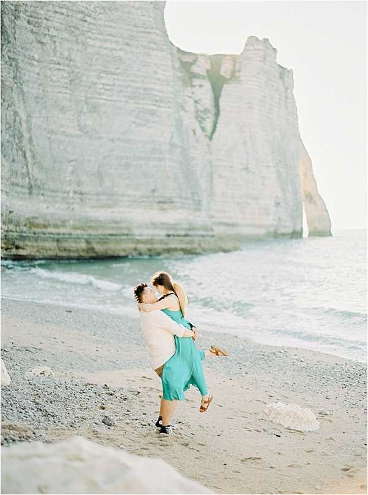 An engagement or couple portrait session on a beach at the base of the famous white chalk cliffs of Étretat in Normandy, France. A man in a cream blazer and black-and-white sneakers lifts a woman wearing a teal jumpsuit and tan sandals, with both leaning in toward each other. The shot is a medium-wide portrait taken at beach level, with the iconic cliff arch visible in the background and gentle waves along the shoreline. The overall color palette is soft and airy, consistent with film photography processing.