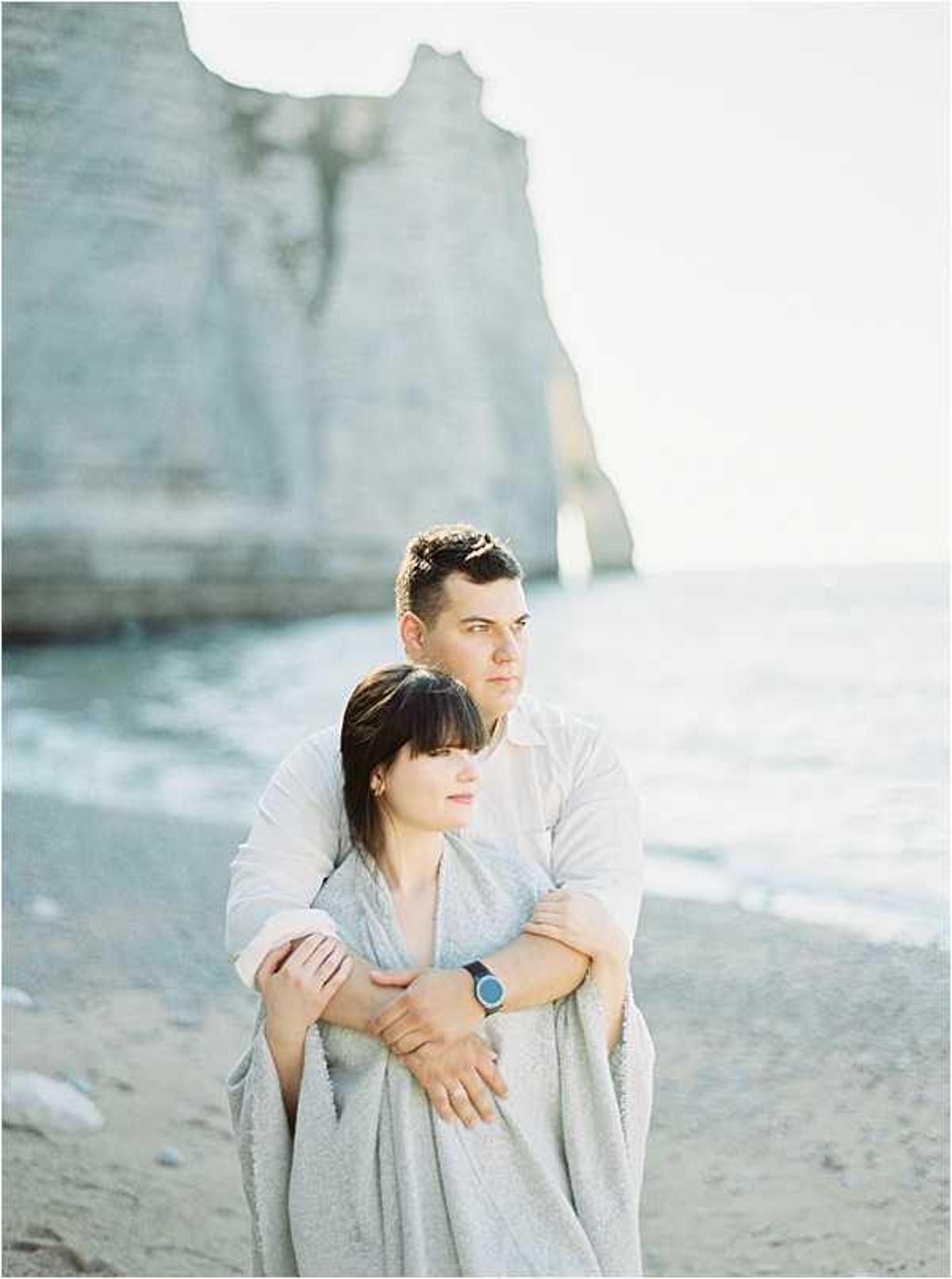 A couple portrait shot on a pebble beach with dramatic white chalk cliffs visible in the soft-focus background, consistent with the cliffs at Étretat in Normandy. The man stands behind the woman with his arms wrapped around her; she is wrapped in a light grey blanket and both look off to one side rather than at the camera. He wears a white linen shirt and a dark-strapped watch, while she wears a light-toned outfit beneath the blanket. The image has a very soft, faded, film-style exposure with cool, pale tones throughout. This appears to be an engagement or pre-wedding couple session rather than a wedding day shot. Medium portrait composition with a shallow depth of field.