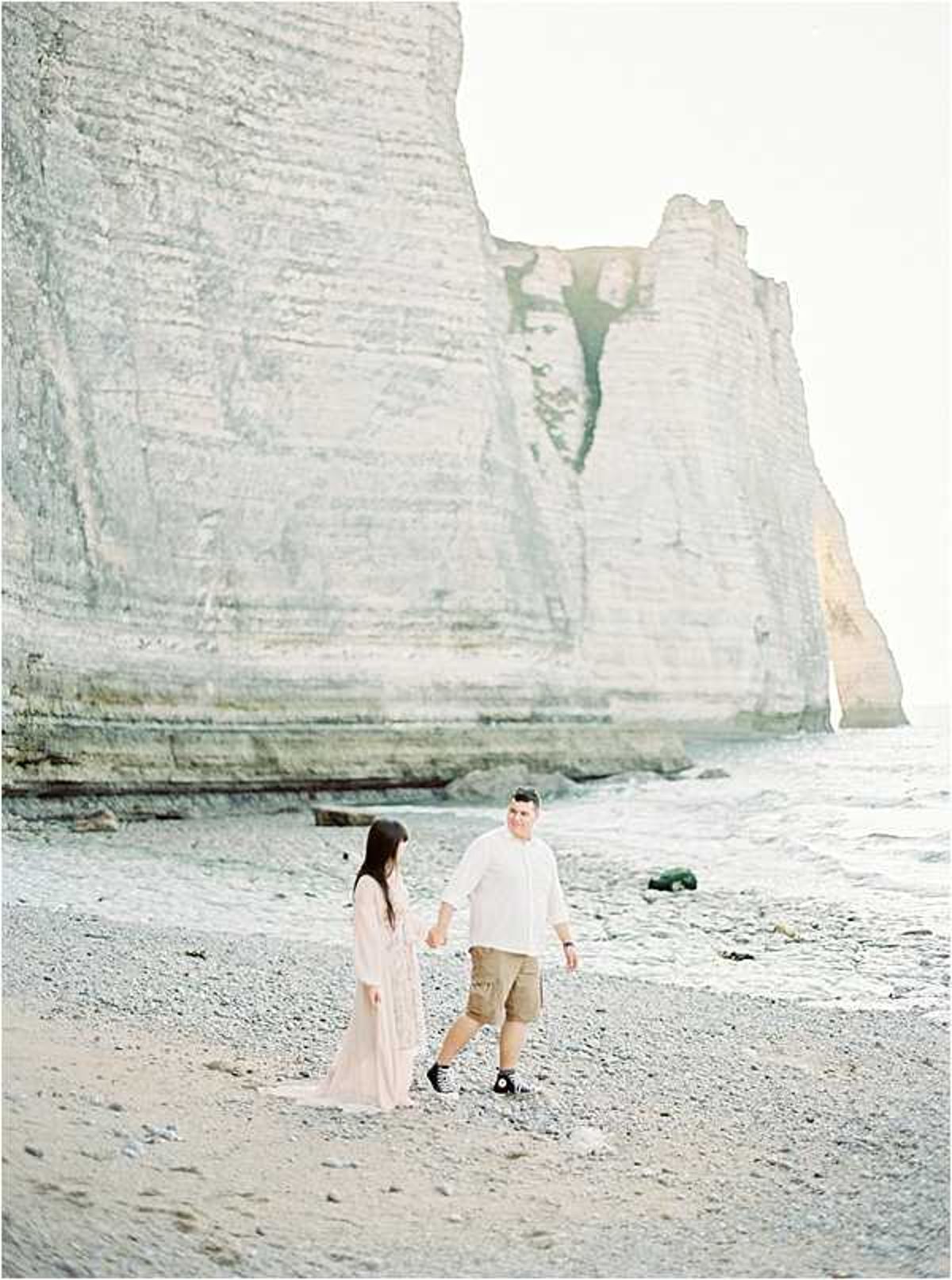 A couple walks hand-in-hand along a pebble beach at the base of tall white chalk cliffs, likely Étretat in Normandy. The woman wears a flowing blush pink maxi dress and the man wears a white linen shirt, khaki shorts, and black-and-white sneakers. The shot is a wide portrait taken in soft, bright natural light with a film-style airy palette. The overall styling is casual and relaxed rather than formal.