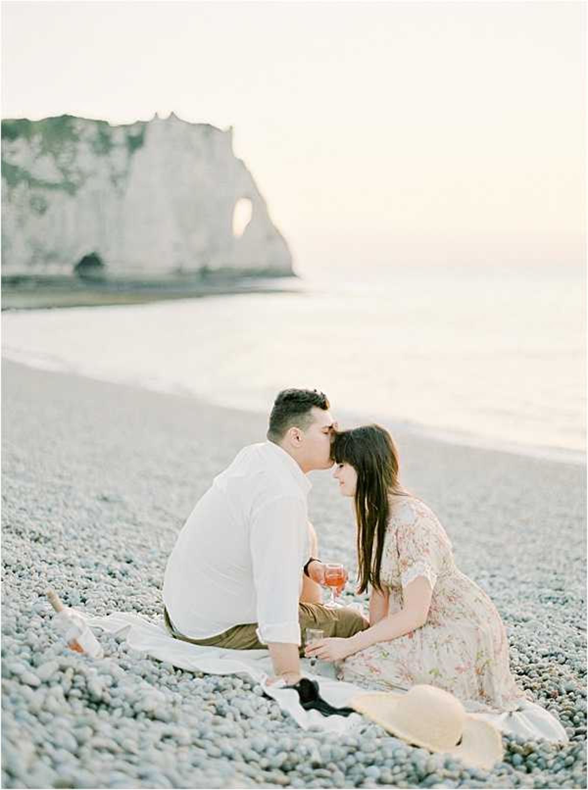 A couple shares a close, near-kissing moment during an engagement or couples portrait session on a pebble beach, identifiable as Étretat on the Normandy coast, with the iconic chalk cliff arch visible in the background. The man wears a white linen shirt and olive trousers, while the woman wears a light floral midi dress with short sleeves; a wide-brim straw hat rests on the pebbles beside them. They are seated on a white blanket, each holding a glass of what appears to be rosé or aperol spritz. The shot is a medium-distance portrait with warm, soft golden-hour light and a pale, washed-out palette throughout.