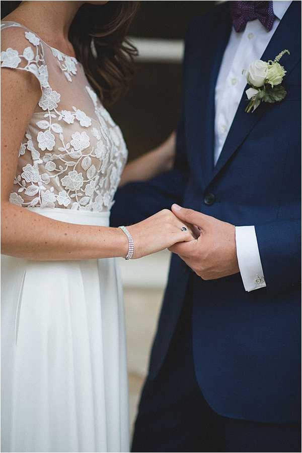 Close-up portrait of a bride and groom holding hands, cropped from shoulders to mid-thigh. The bride wears an ivory gown with a sheer illusion bodice featuring floral lace appliqué and a flowing chiffon skirt, accessorized with a delicate crystal tennis bracelet. The groom wears a navy blue suit with a white dress shirt, burgundy patterned bow tie, silver cufflinks, and a boutonnière of small ivory roses with greenery. The image focuses on their joined hands as a detail shot, with soft natural lighting and a clean, classic styling aesthetic.