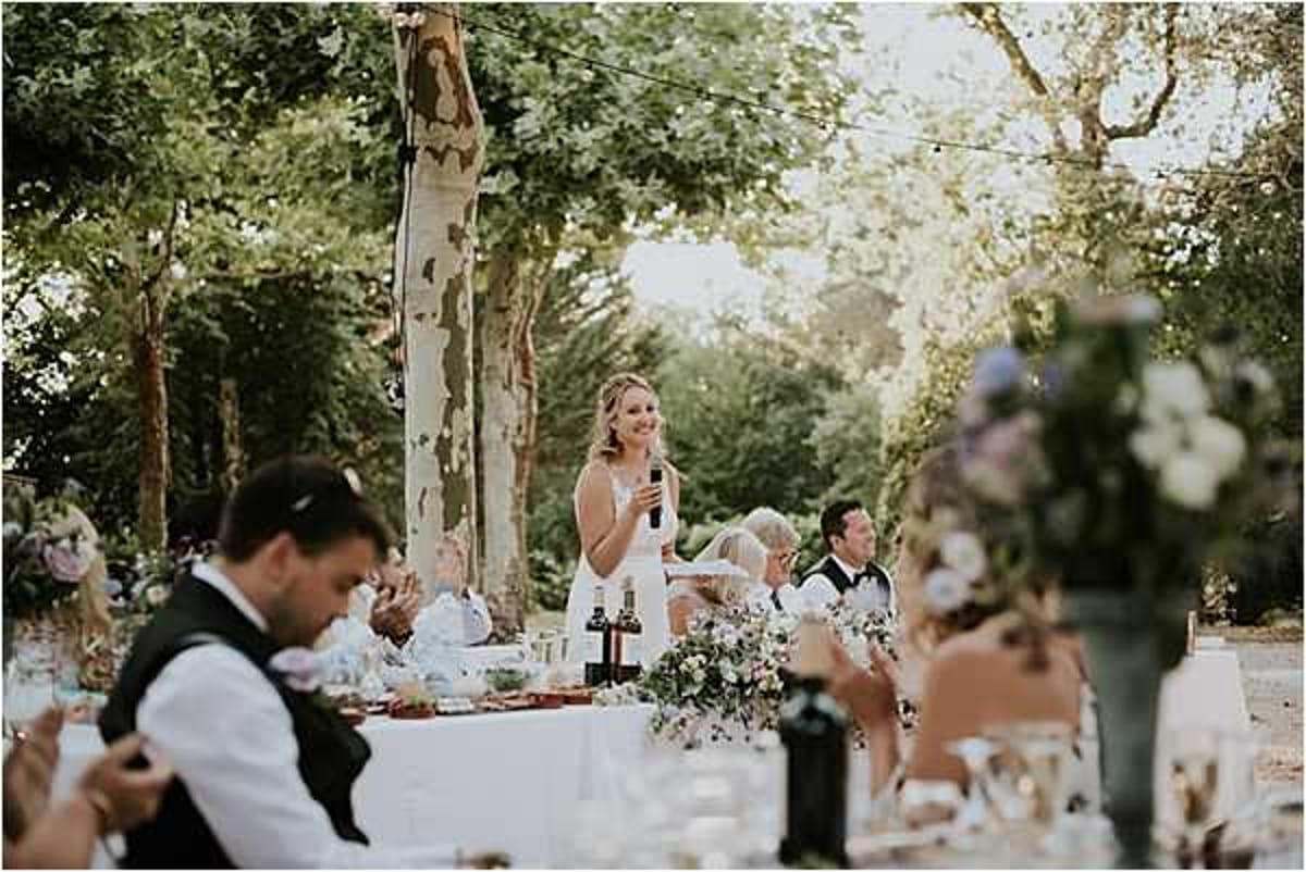 An outdoor wedding reception speech is taking place, with a woman in a white sleeveless dress standing and speaking into a microphone at a long dining table. The setting is a tree-lined outdoor venue with string lights strung between the trees overhead. Approximately 10–15 guests are seated at the table, which is decorated with low floral arrangements featuring soft purple, blue, and white blooms with greenery, along with glassware and wine bottles. In the foreground, a blurred male guest in a white shirt and dark vest is visible, along with an out-of-focus floral centerpiece in similar lavender and blue tones. The overall decor palette is soft and muted, with a relaxed garden-party aesthetic. Wide shot with shallow depth of field.