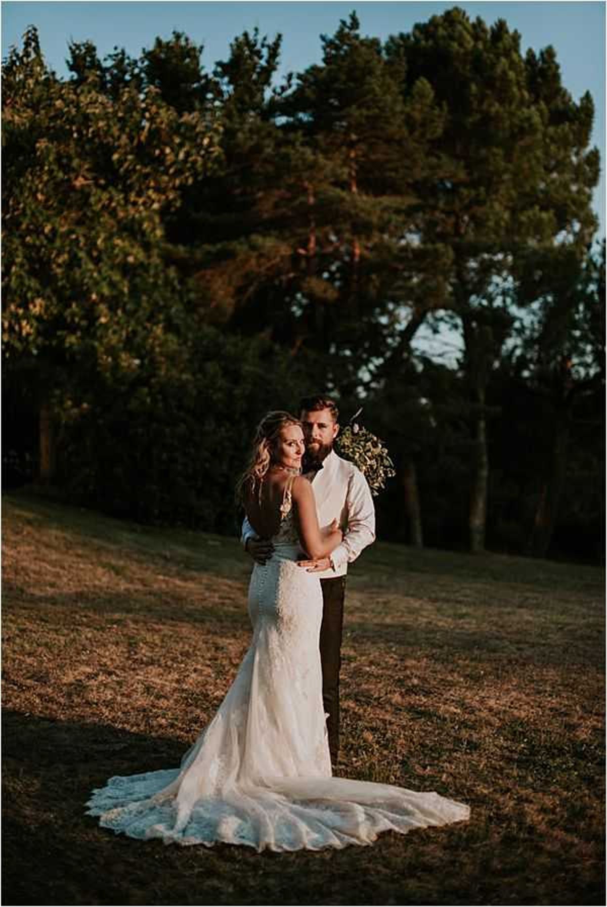 A couple portrait taken outdoors at golden hour, with the bride and groom standing together on an open grassy area with tall trees in the background. The bride wears a fitted, low-back lace gown with a chapel-length train and thin spaghetti straps, with button detailing down the spine; she looks back toward the camera over her shoulder. The groom, bearded, wears a white dress shirt and dark trousers, and holds what appears to be a small greenery-forward bouquet. The warm, low-angled sunlight casts long shadows and a golden tone across the scene, giving the image a moody, rustic feel. This is a full-length couple portrait shot.