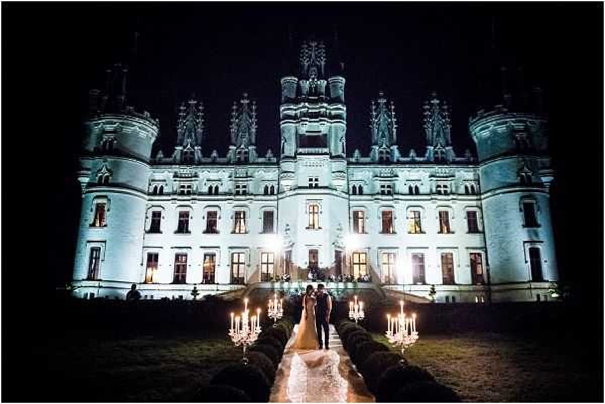 A couple poses outdoors at night on the grounds of a large French Renaissance-style chateau, which is dramatically lit with blue-white uplighting that illuminates its ornate turrets, decorative stonework, and multi-story facade. The bride wears a full-length white gown with a train, and the couple stands at the center of a pathway lined symmetrically on both sides with tall candelabras holding multiple lit candles, creating a warm amber glow along the walkway. The wide-angle shot captures the full scale of the chateau in the background while centering the couple in the foreground, with warm candlelight contrasting against the cool architectural lighting. Potential venue feature image.