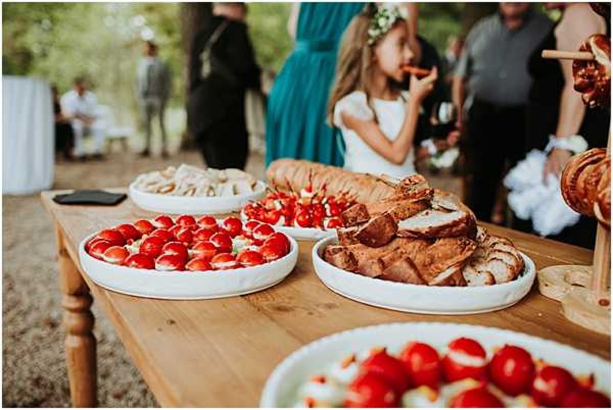 An outdoor cocktail hour food spread displayed on a rustic wooden table, featuring white platters of fresh strawberries, sliced roasted meat, cherry tomato and mozzarella skewers, bread, and additional appetizers. A young flower girl wearing a white dress and a floral crown is eating a snack in the background, while several guests in formal attire — including a woman in a teal dress — mingle in the blurred background. The setting appears to be a garden or wooded outdoor area with a casual, rustic styling. Close-to-medium shot focused on the food table with shallow depth of field.