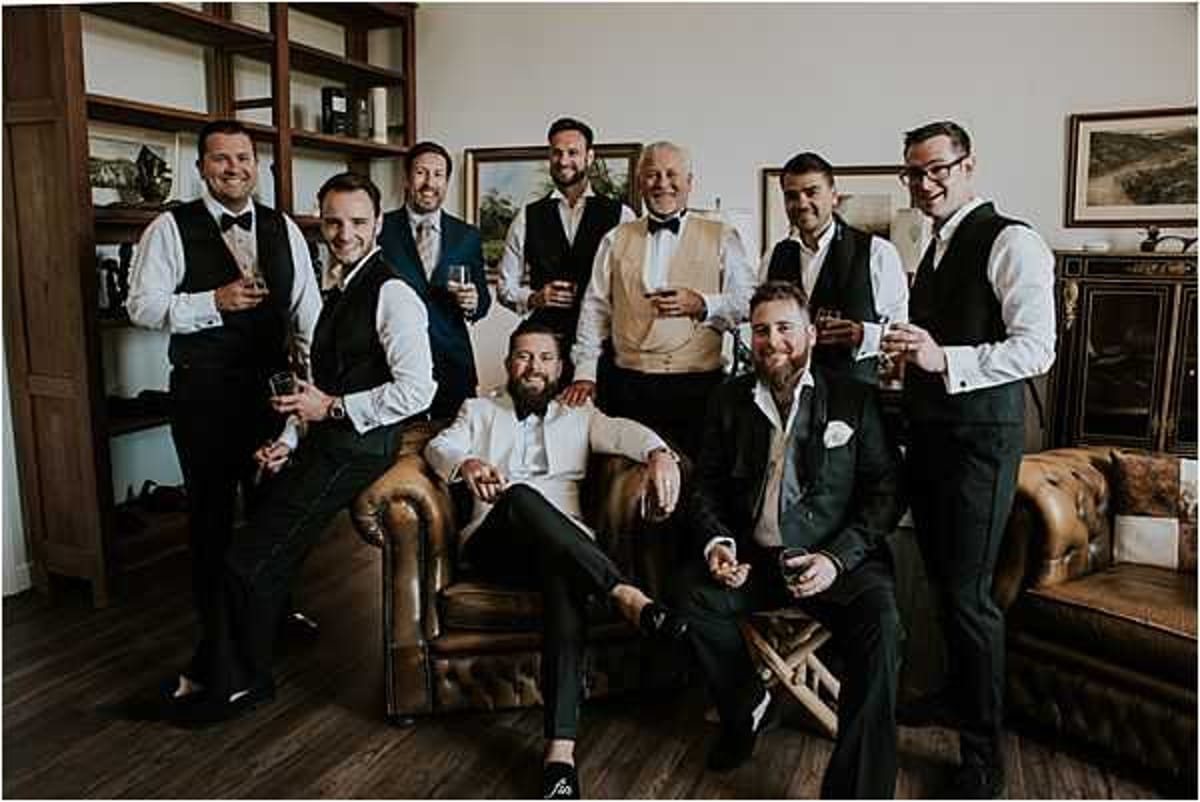 A groom and his groomsmen are posing together in a getting-ready scene indoors, in what appears to be a well-appointed lounge or hotel room with a leather Chesterfield sofa, wooden bookshelves, framed artwork, and a fireplace. The group of nine men includes the groom seated centrally in a white shirt without a waistcoat, while most groomsmen wear dark green or navy waistcoats with white dress shirts, dark trousers, and bow ties or neckties; one older gentleman in the back wears a gold waistcoat. Several men are holding glasses of whisky or champagne. The shot is a wide group portrait with relaxed, candid posing, mixing seated and standing positions around the leather sofa and armchair.