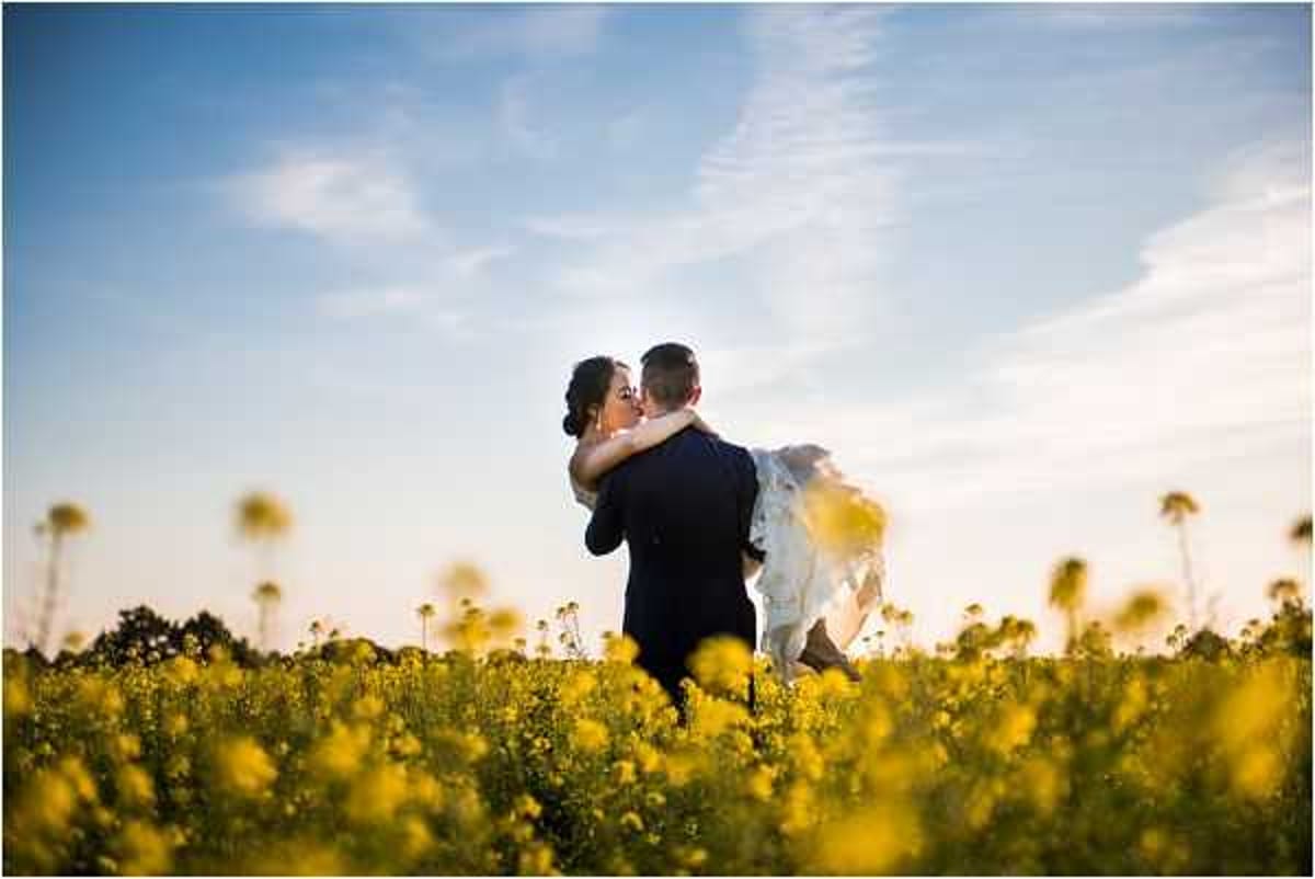 A couple portrait shot outdoors in a field of bright yellow rapeseed flowers during golden hour. The groom, wearing a dark navy suit, is lifting the bride, who is dressed in a white gown, as they kiss. The composition is a wide shot with the couple silhouetted against a pale blue sky in the middle distance, framed by the vivid yellow blooms filling the foreground. The warm evening light creates a soft glow around the couple.
