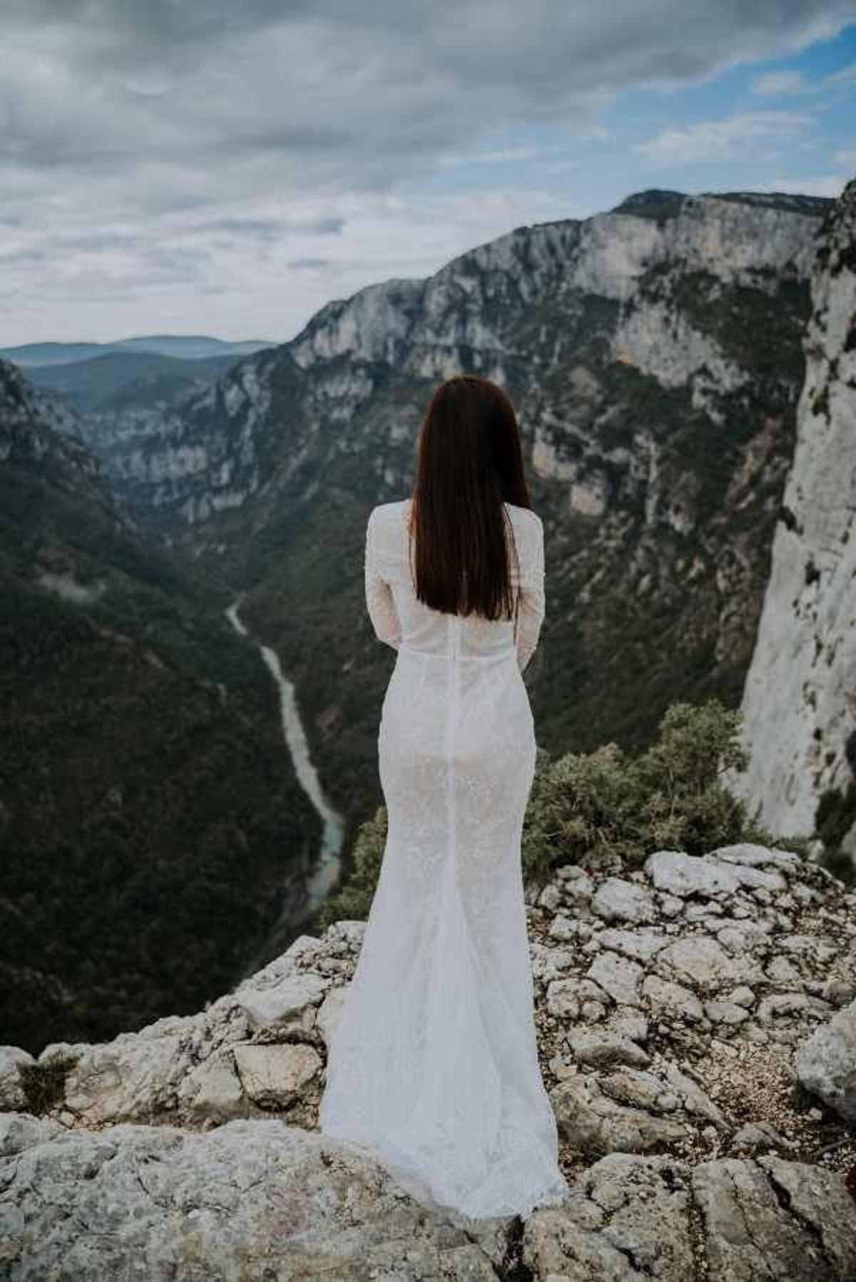 A bride stands alone on a rocky cliff edge, photographed from behind, gazing out over a dramatic canyon with a river winding far below. She wears a fitted, long-sleeve white lace wedding dress with a small train, styled in a boho-romantic aesthetic. Her long dark hair falls loose down her back. The portrait-style shot is taken at cliff-top height, likely at the Gorges du Verdon in southeastern France, and captures a moody, overcast atmosphere.