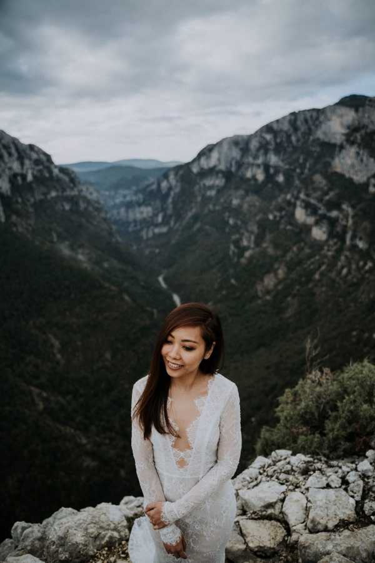 A bride portrait taken outdoors at a dramatic canyon overlook, likely the Gorges du Verdon in southern France. The bride wears a long-sleeve white lace gown with a deep V-neckline and fitted silhouette, and she is looking downward with a slight smile. The composition is a medium portrait shot with the bride positioned in the lower foreground against a wide valley and canyon landscape receding into the distance under an overcast sky.