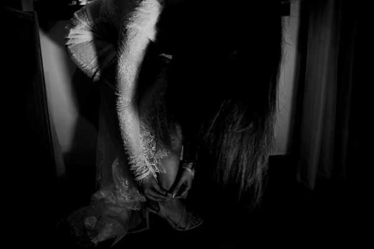 A black-and-white getting-ready shot of a bride bending down to fasten her shoe, captured in a dimly lit indoor room. The image is high-contrast with deep shadows dominating most of the frame, with the bride's lace wedding dress and long dark hair emerging from the darkness. The dress features detailed lace detailing visible on the sleeves and skirt. The composition is a close-up portrait shot taken at a low angle, emphasizing the intimate, candid nature of the moment.