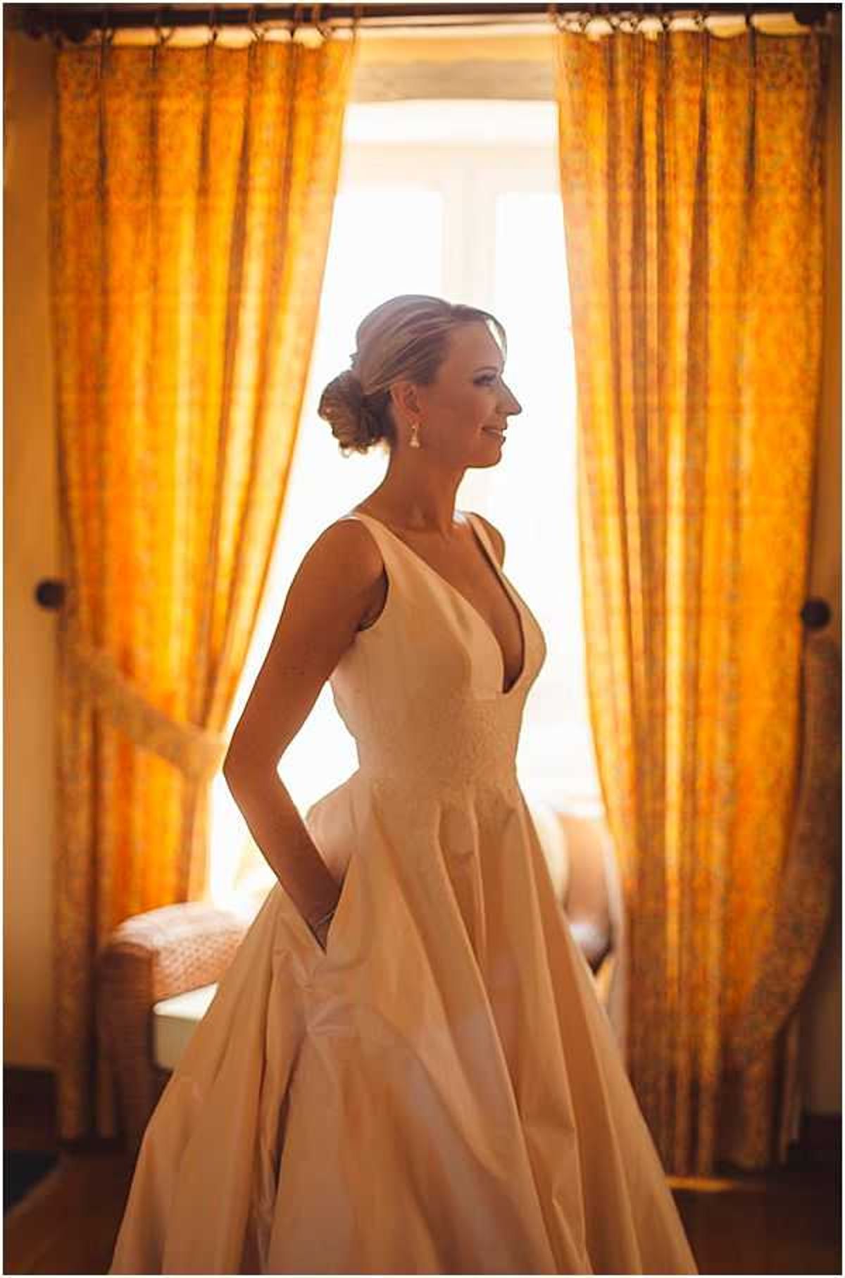 A bridal portrait taken indoors during the getting-ready phase, with the bride standing in front of a large window framed by gold and amber patterned curtains. The bride wears an ivory satin ballgown with a deep V-neckline, wide embroidered waistband, and side pockets, with her hands resting in the pockets. Her hair is styled in a low chignon and she wears drop earrings. The backlit window creates a warm glow around her silhouette, contrasting with the rich golden tones of the curtains and the softly lit interior room behind her. The composition is a three-quarter-length portrait shot taken at a slight angle.