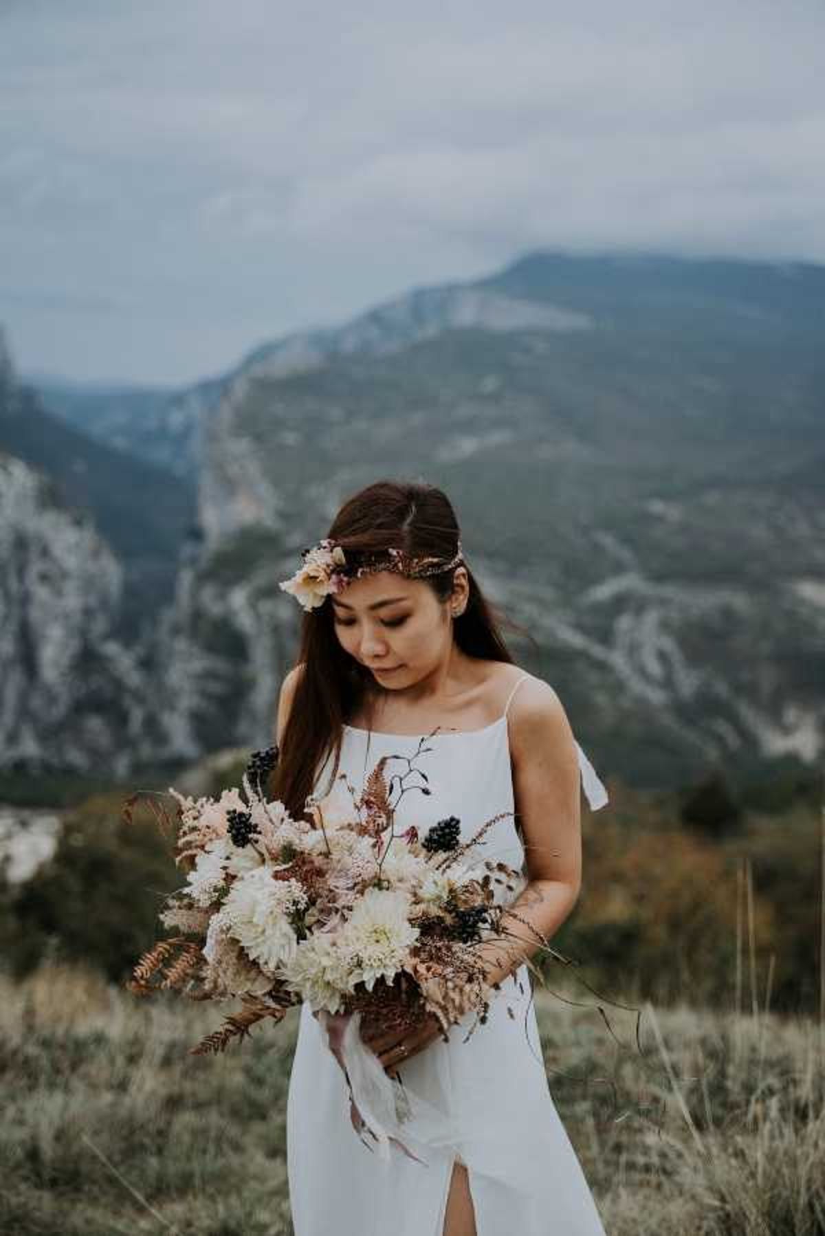 A bridal portrait shot outdoors in a mountainous landscape, with dramatic rocky cliffs and peaks visible in the background. The bride wears a minimalist white slip-style dress with thin spaghetti straps and a front slit, accessorized with a delicate floral and metallic gold headpiece in her long dark hair. She holds a large, loosely arranged bouquet featuring cream dahlias, dried pampas-style grasses, copper-toned ferns, dark burgundy berries, and blush accents, with trailing ribbon streamers. The overall styling aesthetic is boho with earthy, autumnal tones, and the image is a mid-length portrait with a shallow depth of field that softly blurs the mountain backdrop.