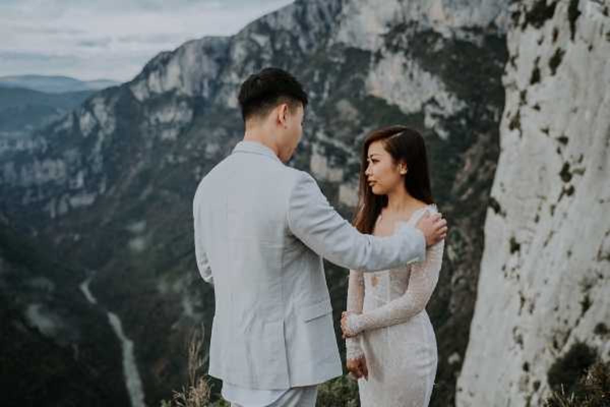 A couple poses for wedding portraits on a rocky cliff edge overlooking a deep gorge and mountain valley, likely the Gorges du Verdon in southern France. The groom, wearing a light grey suit, faces away from the camera and places his hand on the bride's shoulder. The bride faces him wearing a long-sleeved, form-fitting white lace dress with off-shoulder detailing. The composition is a medium portrait shot taken at overcast natural light, with the dramatic canyon and layered mountain ridgeline visible in the background.