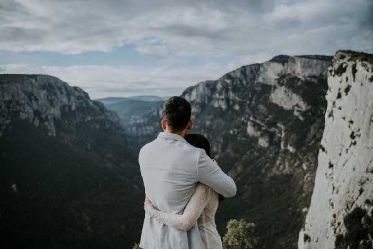 A couple embraces while standing at the edge of a dramatic canyon overlook, viewed from behind in a wide portrait shot. The groom wears a light grey suit jacket and the bride wears a white lace long-sleeve dress, both facing away from the camera toward the expansive gorge and layered mountain ridges in the distance. The setting appears to be the Gorges du Verdon or a similar southern French canyon location, shot outdoors under an overcast sky. The composition emphasizes the scale of the landscape against the two figures, with a moody, muted color palette consistent with an adventurous, nature-forward wedding style.