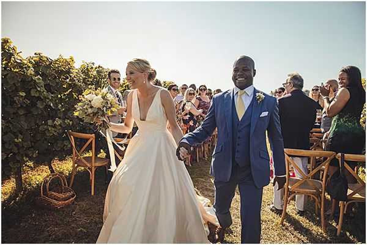 A bride and groom walk back up the aisle together immediately after an outdoor ceremony set within a vineyard, with rows of grapevines visible on either side. The bride wears a deep V-neck, full-skirted ivory gown and carries a loose bouquet of cream and white blooms with greenery, while the groom wears a navy three-piece suit with a light gold tie and a boutonniere. Roughly 30–40 guests seated on cross-back wooden chairs line the aisle, many smiling and applauding. The shot is a wide candid portrait taken at ground level in bright natural sunlight with warm golden tones.