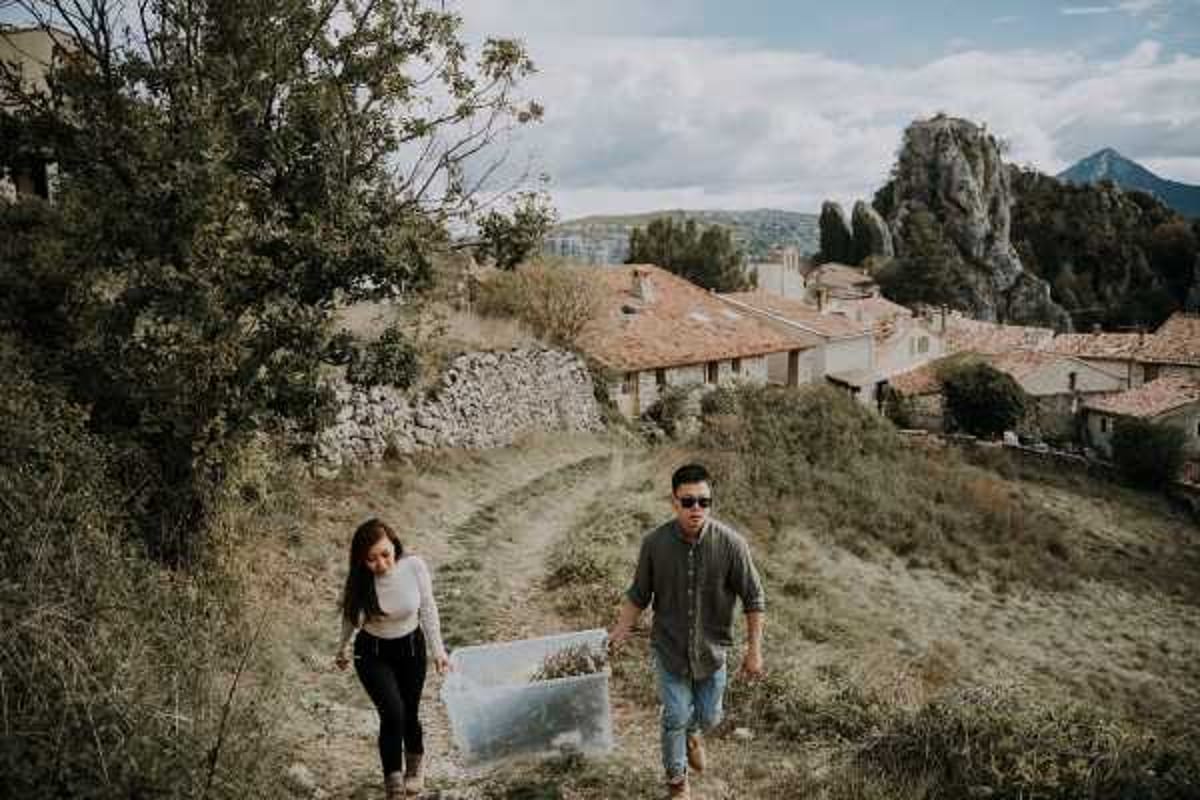 A couple walks up a hillside path together in what appears to be an engagement or pre-wedding portrait session in a French village. The man wears a dark olive button-up shirt and jeans with sunglasses, while the woman wears a white long-sleeve top and black pants; one of them is carrying a clear acrylic box, likely a prop. The setting is an elevated outdoor location overlooking terracotta-roofed stone village buildings, with rocky outcrops and hills visible in the background. The shot is a medium wide portrait taken at ground level, captured in a relaxed, candid documentary style.