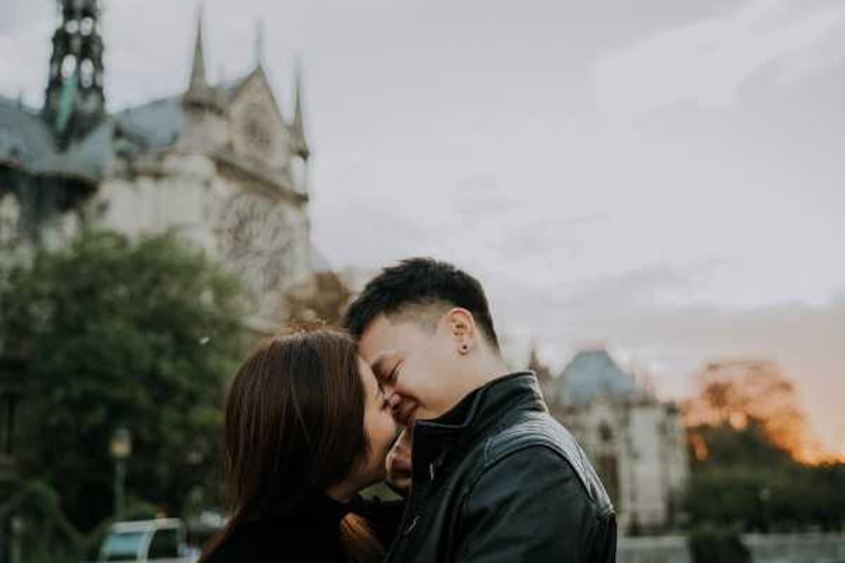 A couple shares a close, intimate moment outdoors, their faces nearly touching in what appears to be a kiss or near-kiss, both smiling with eyes closed. The shot is a close-up portrait with the iconic Notre-Dame de Paris cathedral visible and softly blurred in the background, along with warm golden-hour light on the horizon. The groom wears a black leather jacket, and the bride has long dark hair. The overall styling is casual and relaxed rather than formally bridal, suggesting this may be an engagement or pre-wedding portrait session rather than a wedding day shot.
