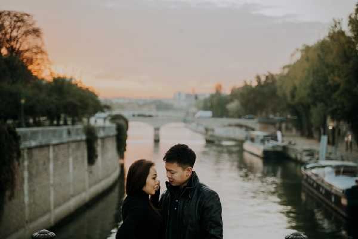 A couple stands close together facing each other along the banks of the Seine River in Paris, captured during a portrait session at sunset. The man wears a dark leather jacket and the woman wears a dark top, both in casual attire suggesting an engagement or pre-wedding shoot rather than a ceremony day. The background shows the Seine with bridges, moored boats, and city architecture visible in the soft warm light of dusk. The composition is a medium portrait shot with the river and Parisian cityscape providing depth behind the subjects.