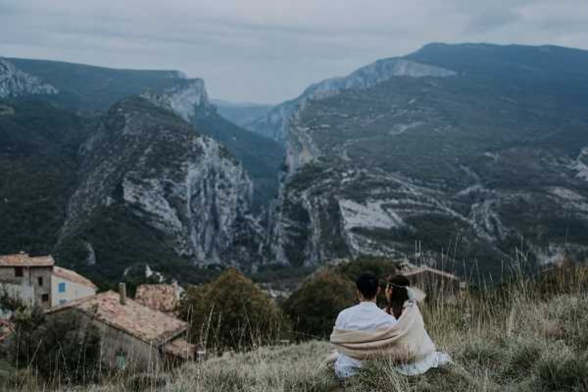 A couple sits together on a hillside overlooking a dramatic canyon landscape with steep rocky cliffs, photographed from behind in a wide shot. The bride wears a white dress and has a floral hair accessory, while both are wrapped in a shared warm beige/cream blanket or shawl. A small stone-roofed village building is visible in the lower left corner of the frame. The styling is boho in feel, with the relaxed seated pose and natural textile wrap adding an informal, intimate quality to the portrait session.