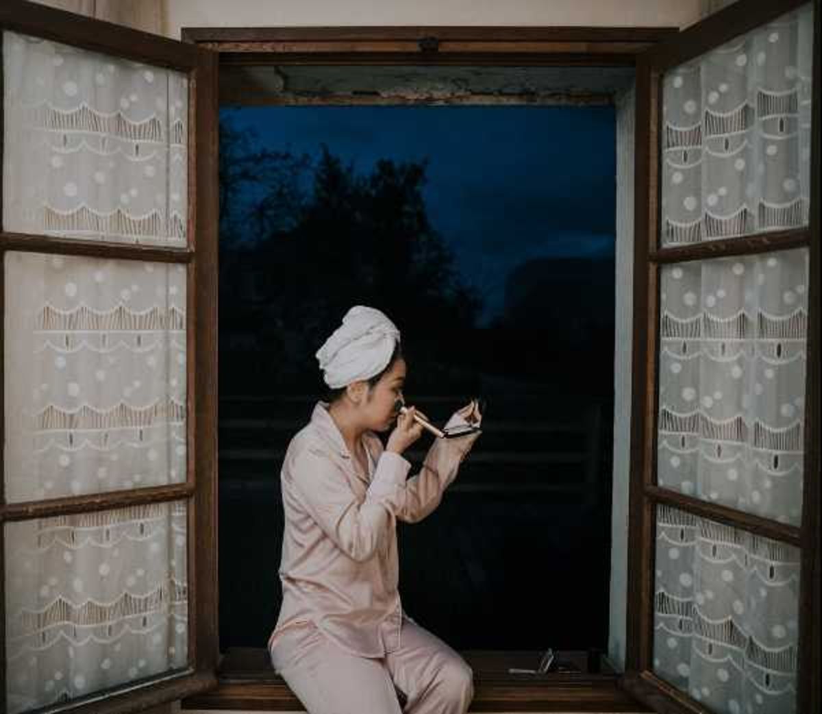 A bride is captured during her getting-ready preparations, seated on a windowsill applying makeup using a small compact mirror. She is wearing blush pink satin pajamas with a white towel wrapped around her hair. The window is framed by wooden shutters and white lace curtains with a polka dot and scalloped pattern. The scene is lit by interior warm light contrasting against a dark blue early-morning or evening exterior visible through the open window. The composition is a medium portrait shot taken from a slight distance, framing the subject centrally within the open window.