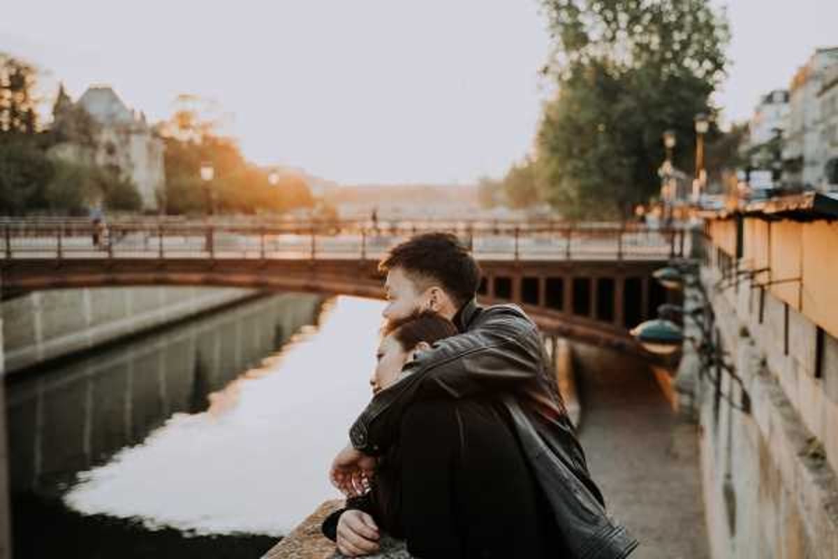 A couple portrait shot outdoors in an urban setting, with the pair standing on a bridge or embankment wall overlooking a canal or river channel. The man, wearing a dark leather jacket, stands behind the woman and wraps his arms around her as both look out over the water. The image is taken at golden hour, with warm backlit sunlight creating a soft glow across the scene. The composition is a medium portrait shot from the side, with city buildings, a bridge, and trees softly out of focus in the background.
