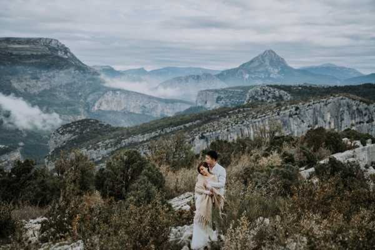 A couple portrait shot outdoors in a dramatic mountain landscape, likely the Gorges du Verdon region of France. The bride wears a white gown and is wrapped in a cream or beige fringed shawl, held close by the groom who is dressed in a white shirt. The two stand among dry golden shrubs and rocky terrain, embracing and facing each other intimately. The wide shot composition places the couple in the lower-middle third of the frame, with layered mountain ridges, canyon cliffs, and low clouds filling the expansive background. The overall styling is relaxed and bohemian, with the shawl serving as both a practical and aesthetic detail against the cool, overcast conditions.