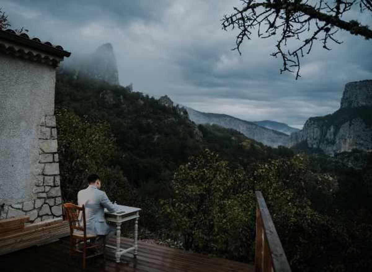 A groom in a light grey suit sits alone at a small white desk and wooden chair on an outdoor wooden terrace, appearing to write or review notes — likely wedding vows. The terrace is attached to a stone building and overlooks a dramatic mountain gorge landscape under overcast, moody skies. The wide-shot composition places the solitary figure small against the vast backdrop, emphasizing the scale of the setting. The overall palette is cool and muted, with deep blue-grey tones throughout the image.