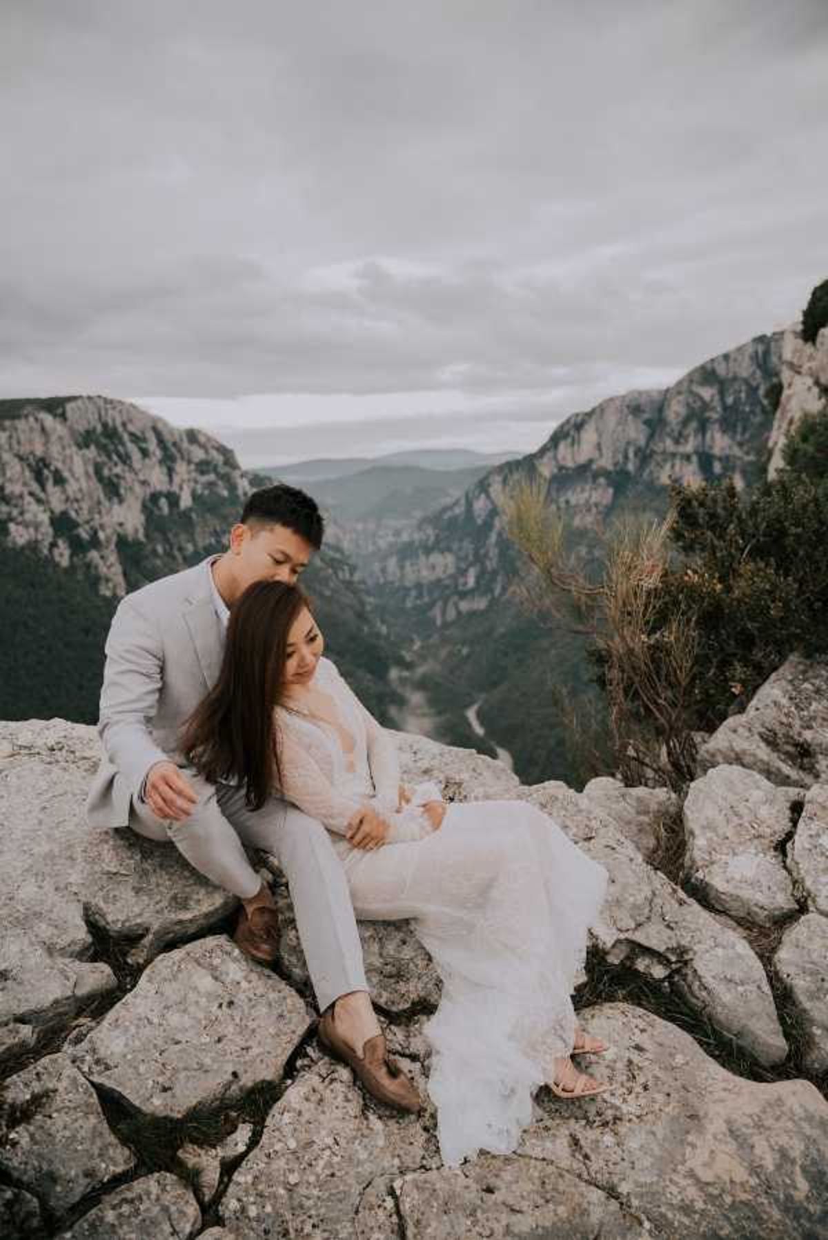 A couple portrait shot outdoors at a dramatic clifftop overlook, with a deep canyon and winding river visible in the background, consistent with the Gorges du Verdon in southern France. The groom is dressed in a light grey suit with brown leather loafers, and the bride wears a long-sleeve lace wedding gown with a deep V-neckline and a fitted silhouette with a trailing skirt. The two are seated closely together on a rocky ledge, with the groom looking down toward the bride, who is smiling softly downward. The overall styling is boho-romantic, and the overcast sky gives the image a moody, muted tone. This is a medium-distance couple portrait with a wide landscape background.