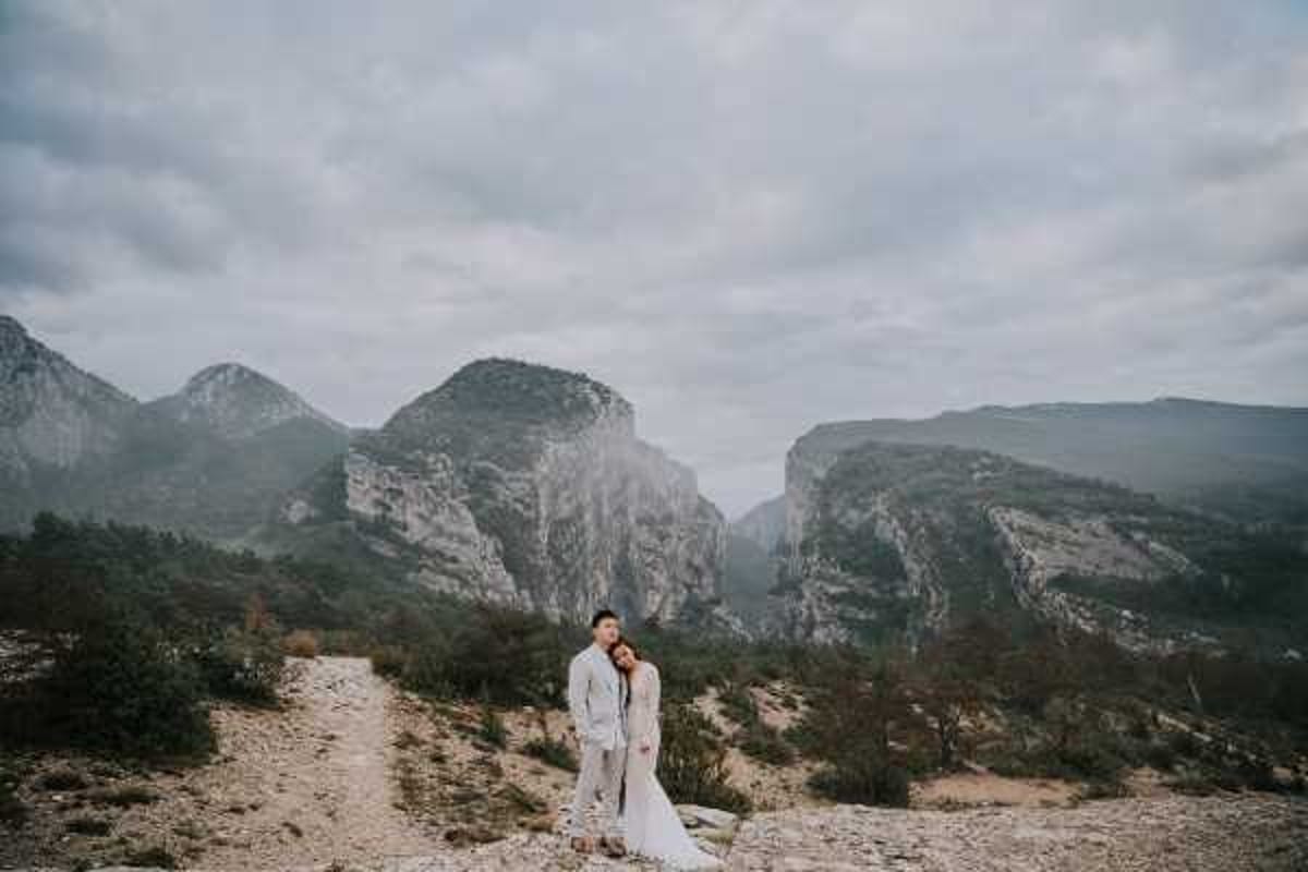 A couple poses together on a rocky outdoor overlook with dramatic canyon cliffs and misty mountains in the background, in what appears to be the Gorges du Verdon region of France. The bride wears a long-sleeved white lace gown with a train, and the groom is dressed in a light grey suit. They stand close together on a pale rocky path, the bride leaning into the groom. This is a wide environmental portrait shot that places the couple small within the vast landscape, emphasizing the scale of the surrounding terrain.