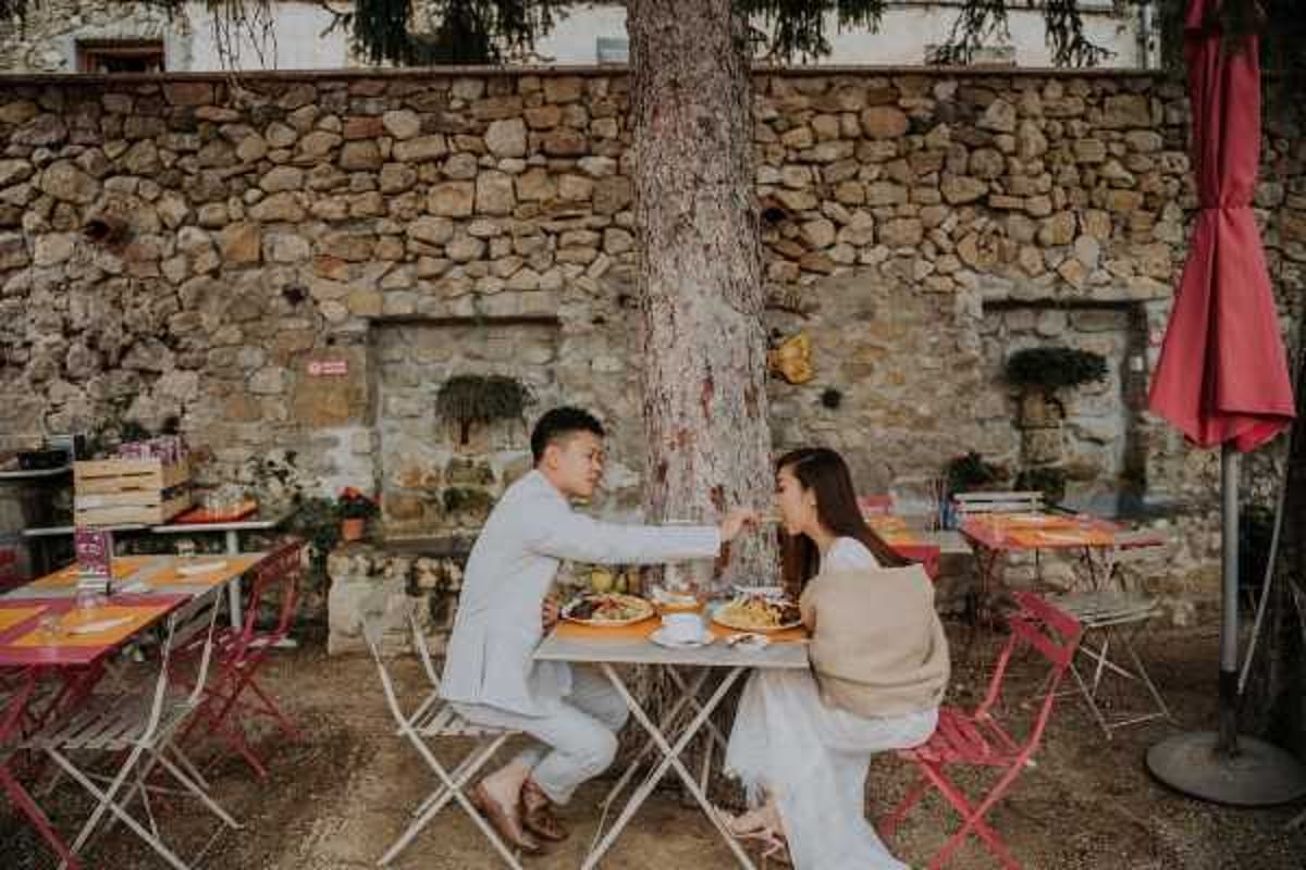 A couple shares a casual outdoor meal at a small white bistro-style folding table set against a rustic stone wall, with a large tree trunk at the center of the frame. The man wears a light grey suit and the woman wears a white dress with a beige wrap draped over her shoulders. Their table is set with plates of food and drinks, and the surrounding terrace features coral-red metal bistro chairs and tables. The shot is a medium wide portrait capturing an candid, relaxed moment between the two, styled in a casual French café setting.