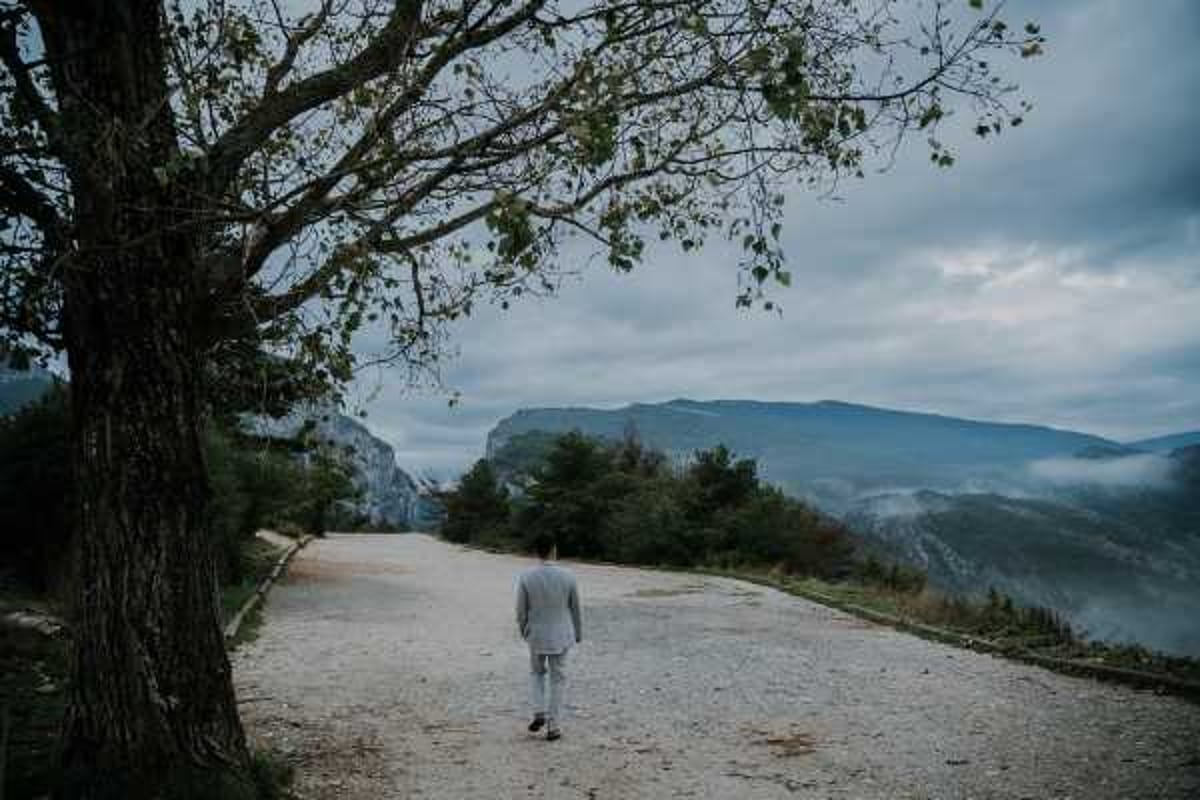 A groom walks alone down a wide gravel path toward a dramatic mountain landscape, shot from behind in a wide environmental portrait. He is wearing a light grey suit and is positioned centrally in the frame, dwarfed by the expansive scenery of rocky cliff faces and misty valley below. A large bare tree frames the left side of the composition. The overall mood is moody and cinematic, with overcast skies and low cloud cover settling between the mountain ridges.