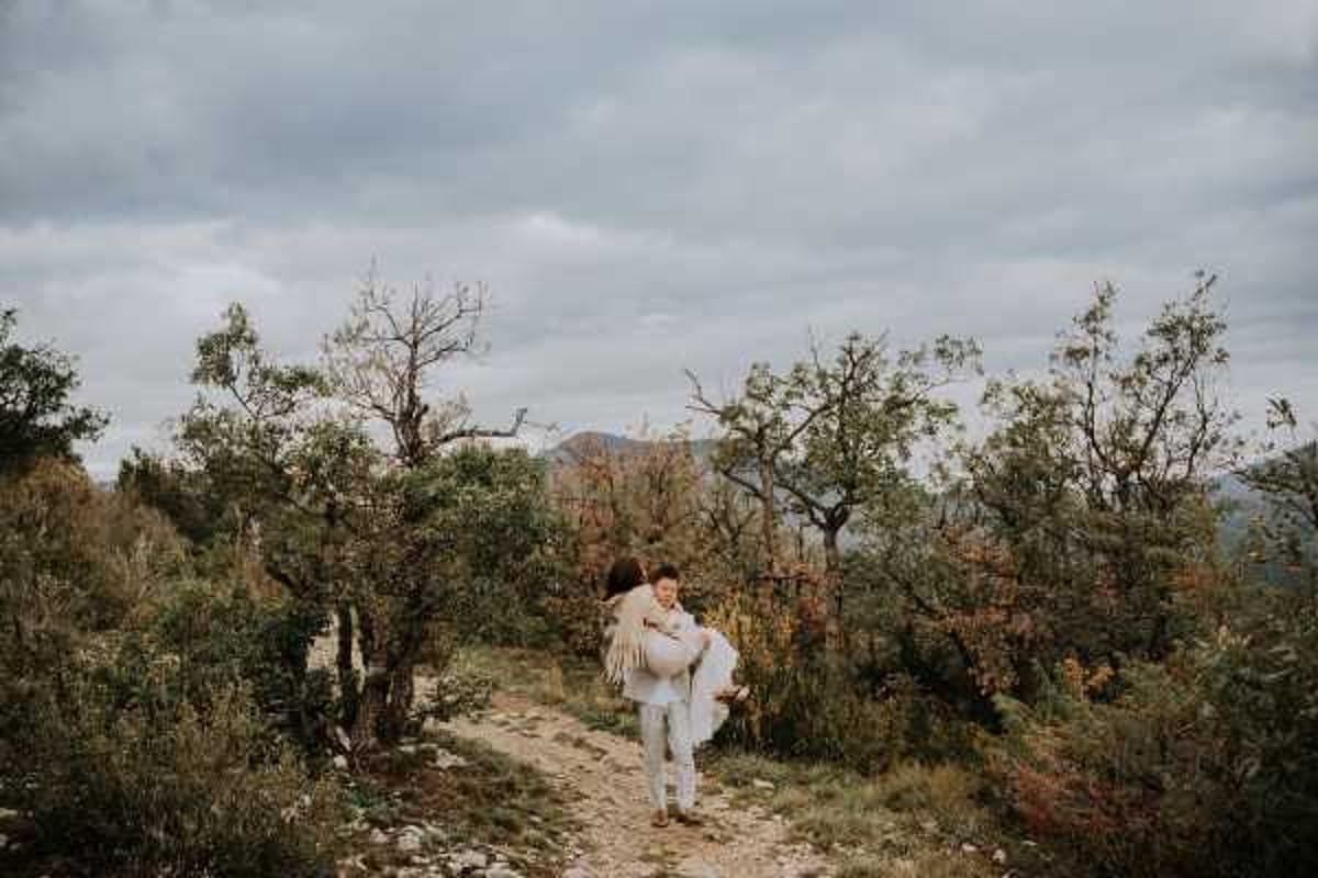 A couple portrait taken outdoors on a rocky hillside trail surrounded by low scrubland and sparse trees, with mountains visible in the background under an overcast sky. The groom, dressed in light grey trousers and a white shirt, is carrying the bride on his back; she wears a white dress with a fringed ivory or cream shawl wrap. The styling has a relaxed boho aesthetic. The shot is a wide environmental portrait capturing the full landscape context around the couple.