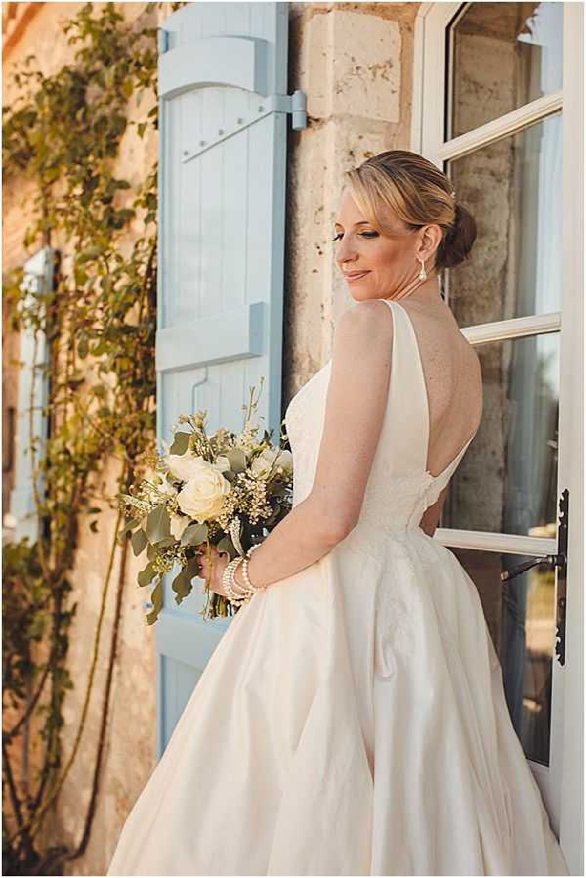 A bridal portrait taken outdoors against the facade of a French stone building featuring pale blue wooden shutters and a white-framed window. The bride stands in three-quarter profile, glancing back over her shoulder toward the camera, wearing an ivory ball gown with a low open back and full skirt. Her hair is styled in a low chignon, and she wears drop earrings and pearl bracelets. She holds a bouquet of cream garden roses, eucalyptus, and small white filler flowers with trailing greenery. The warm golden light suggests late afternoon or early evening. The composition is a medium portrait shot with a shallow depth of field, keeping the bride sharp against the softly blurred architectural background.