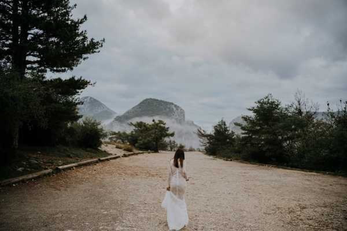 A bridal portrait taken outdoors on a wide gravel path or plateau, with the bride walking away from the camera toward a mountainous landscape partially obscured by low cloud or mist. The bride is wearing a white lace long-sleeve gown with a flowing skirt, her dark hair down. The setting appears to be a rugged, elevated outdoor location with conifer trees on either side of the path. The image is a wide shot with a moody, overcast atmosphere and a boho editorial style.