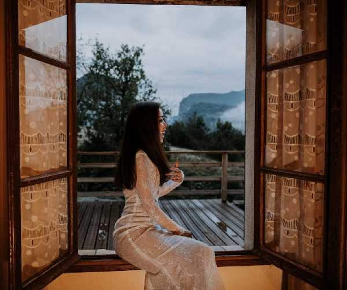 A bride with long dark hair sits in the open frame of a wooden double window, looking out to the side in a relaxed, contemplative pose. She is wearing a long-sleeved, form-fitting ivory lace gown. The window has sheer patterned curtains with a warm golden tone, and opens onto a wooden terrace with a mountainous, misty landscape in the background. The shot is a medium portrait framed by the window architecture, with warm interior tones contrasting against the cool blue-grey outdoor light.