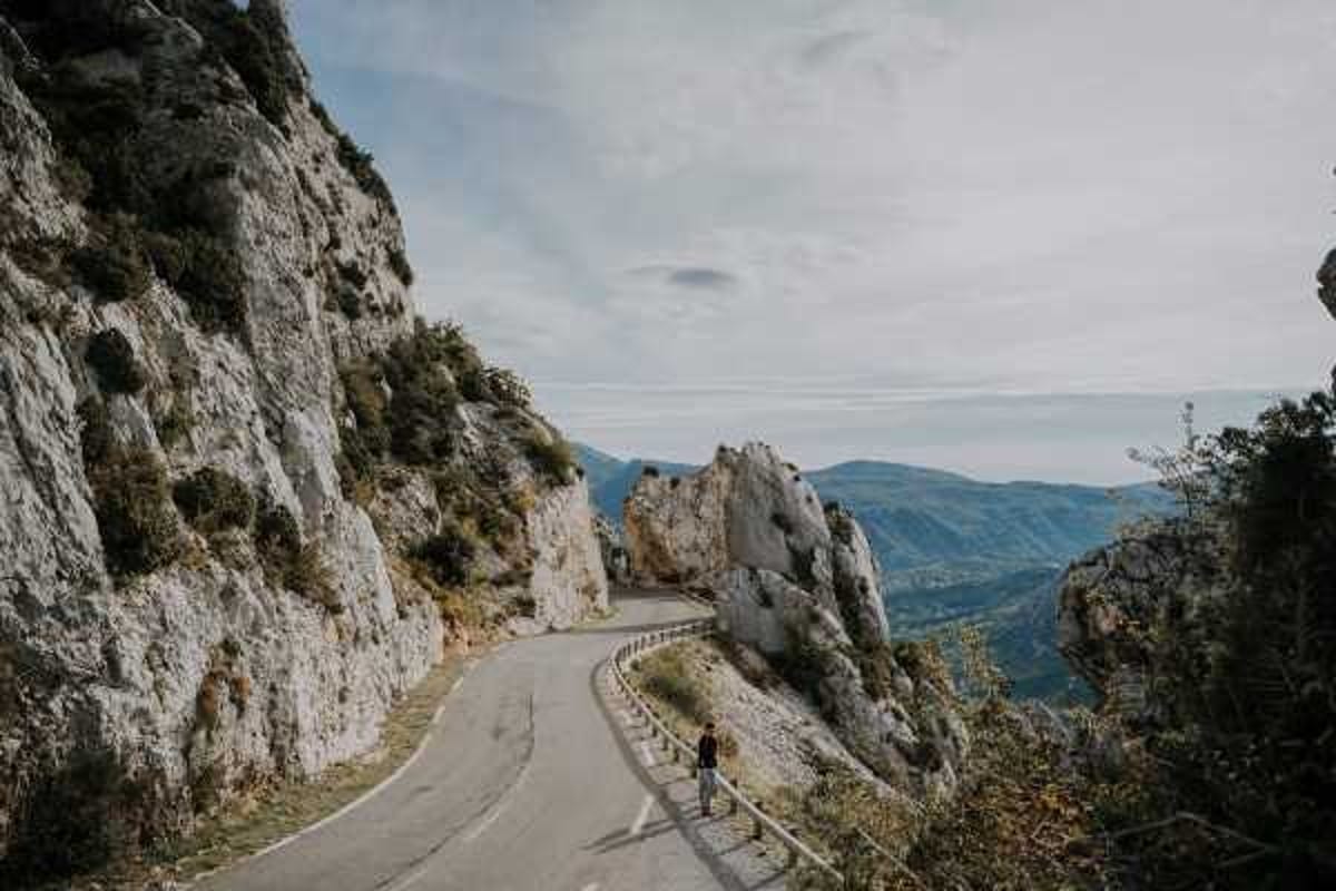 A wide landscape shot showing a single person standing on a narrow mountain road that winds through rocky terrain with a sweeping valley view in the background. The figure appears small against the dramatic scale of the rocky cliffs and mountain scenery, suggesting this may be a couple's portrait session or editorial shot taken on location in a mountainous region of France, possibly the Alps or Verdon Gorge area. The image is shot in a documentary wide-angle style with muted, natural tones. No wedding-specific details such as attire, florals, or decor are clearly visible at this distance.